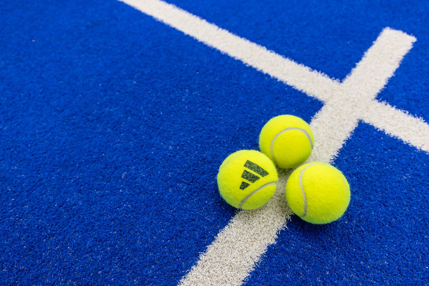 Three yellow Adidas tennis balls stacked on a blue padel court near white boundary lines.