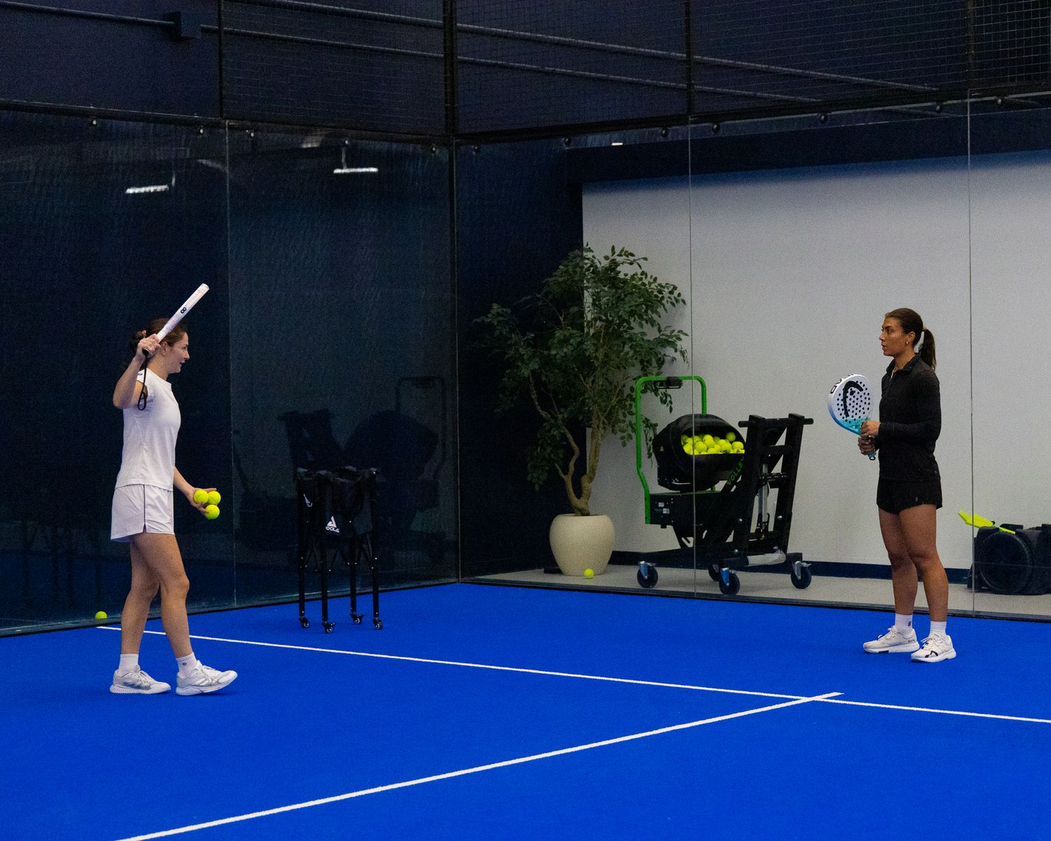 Two women practicing padel tennis on an indoor blue court during a training session.
