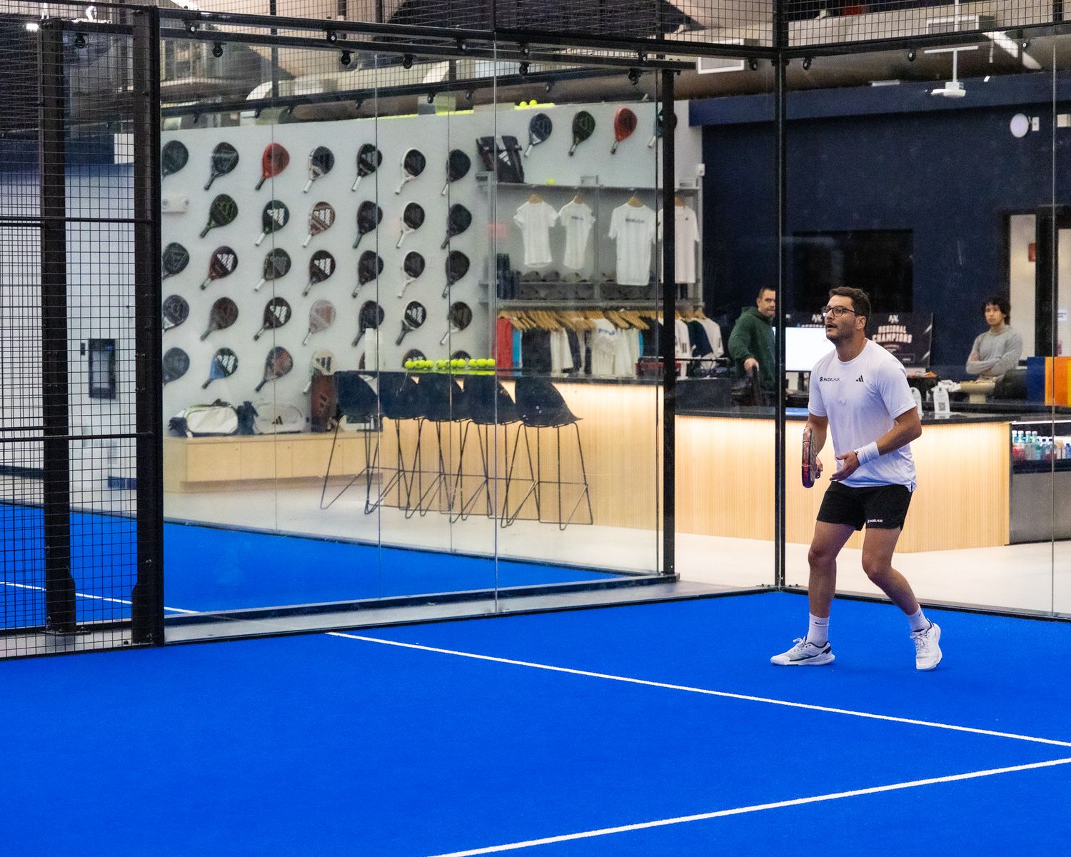 Man playing padel tennis on a blue indoor court with a pro shop visible behind glass.