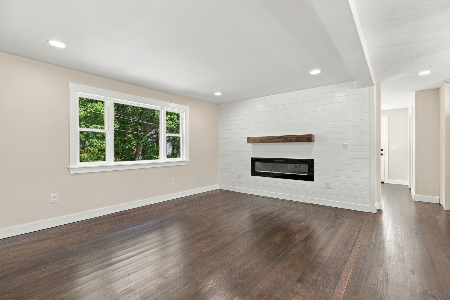 Bright living room with dark hardwood floors, shiplap fireplace wall, recessed lighting, and large window with garden views.