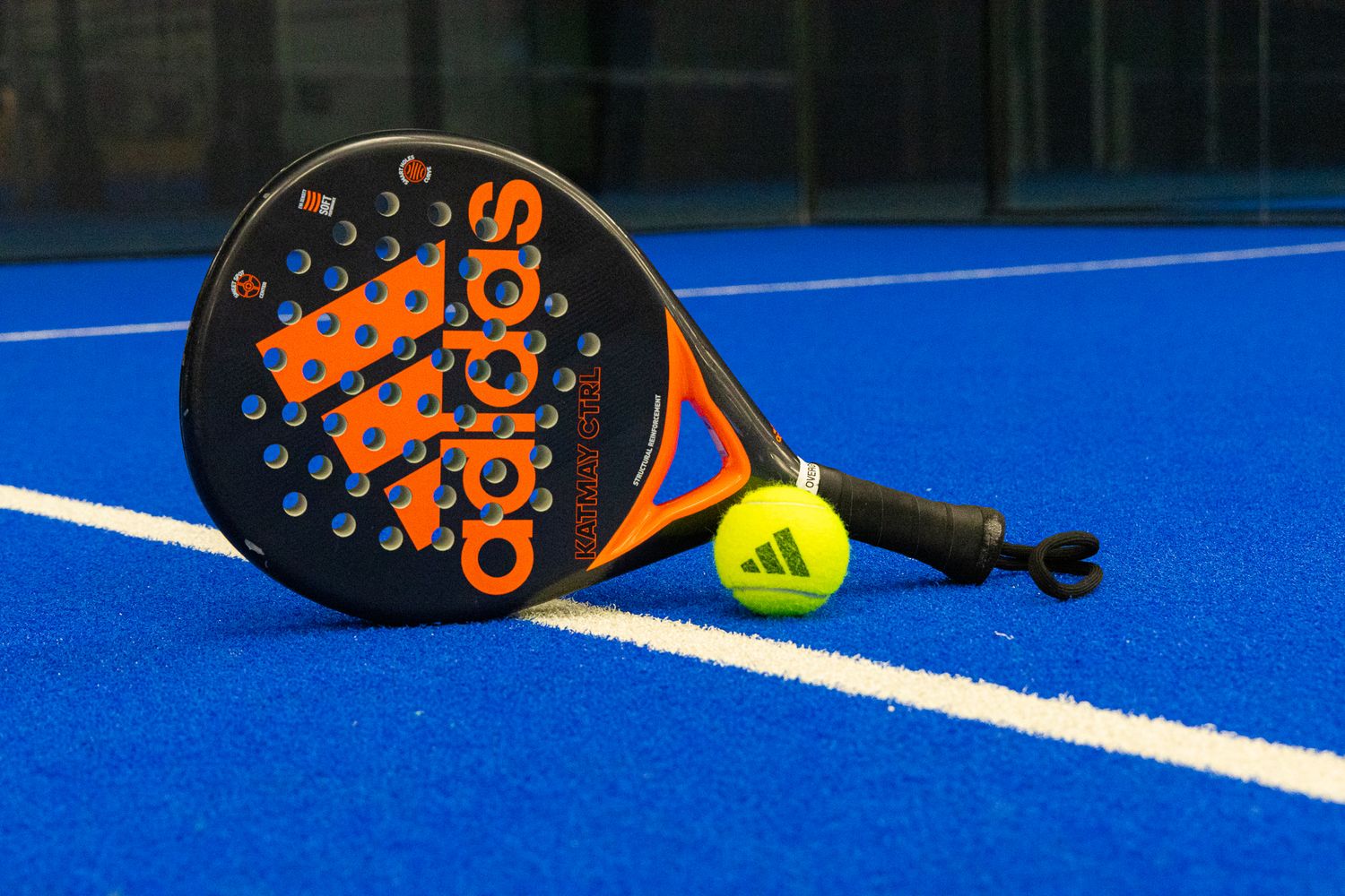 Adidas orange padel racket and yellow ball placed on blue padel court surface.