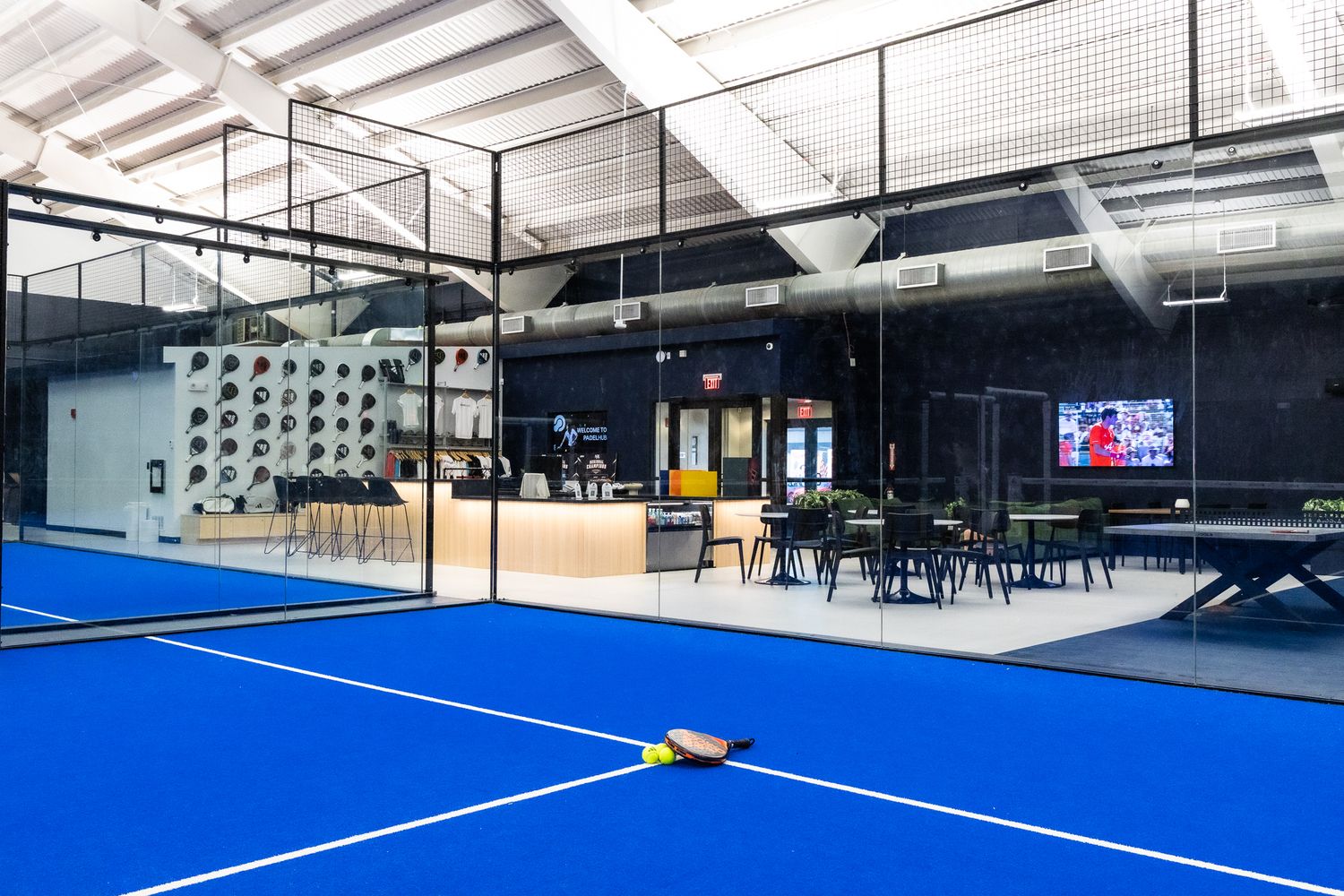 Indoor PadelHub facility showing blue courts, lounge area, TV screen, and seating.