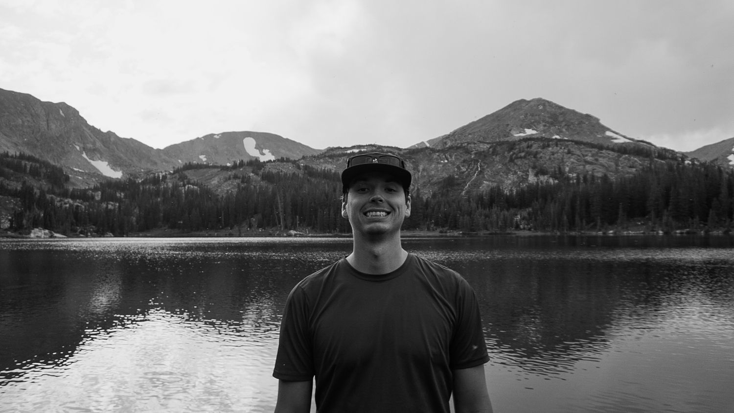 Man smiling by a mountain lake with forested peaks in background, black and white outdoor portrait.