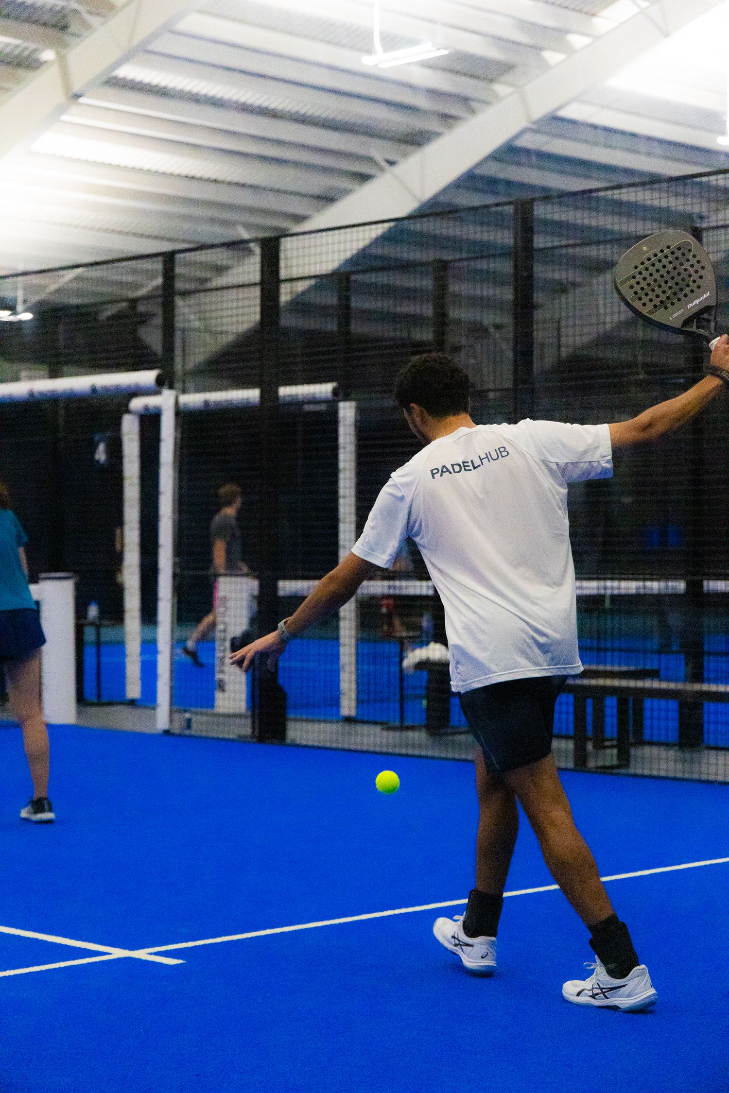 PadelHub branded player hitting a forehand shot on a blue indoor padel tennis court.