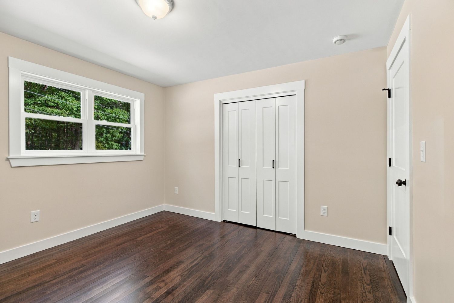 Bright bedroom with dark hardwood floors, white closet doors, and a window with natural light in a neutral-toned room.
