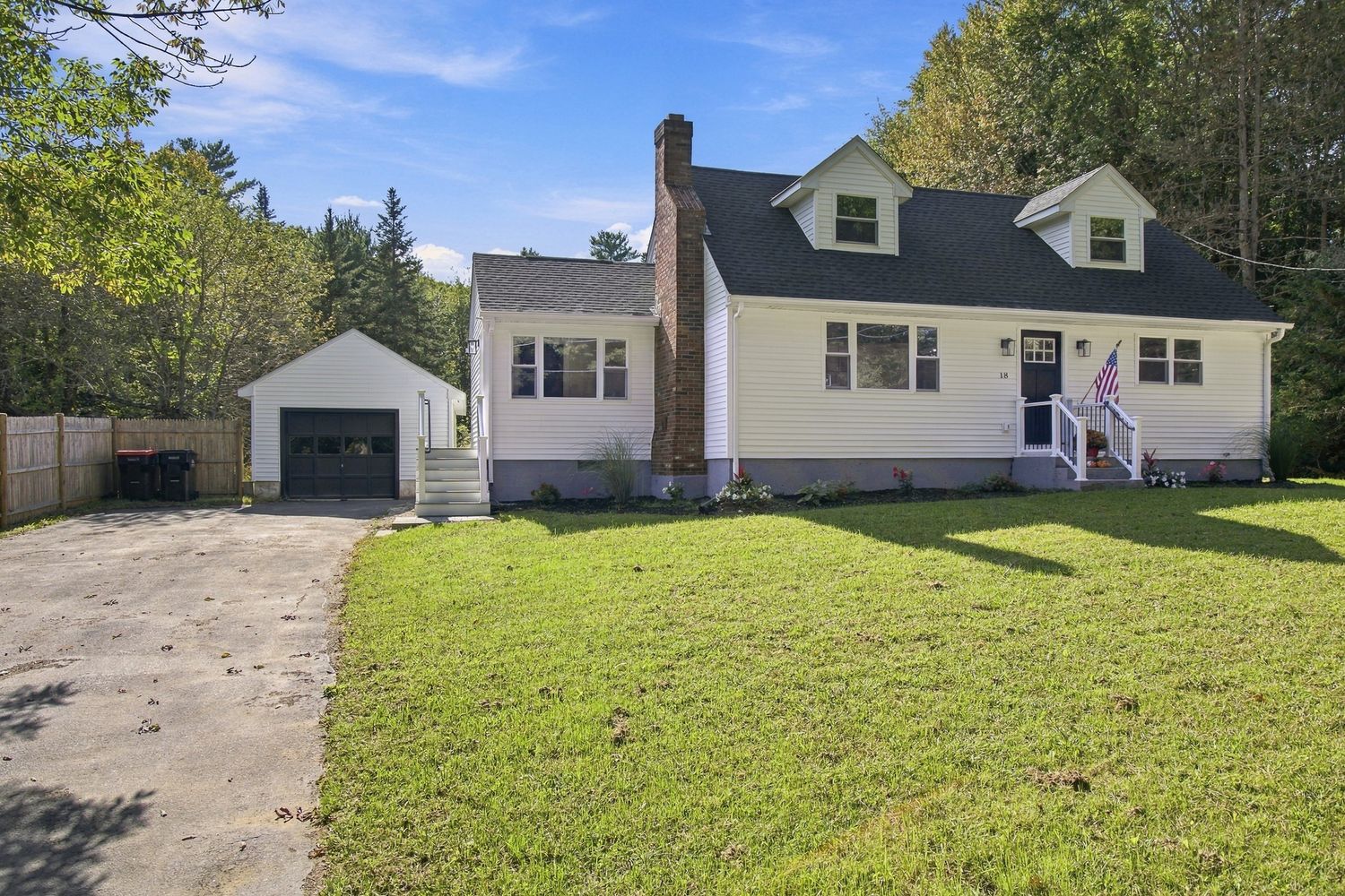 White Cape Cod home with attached garage, green lawn, and tree-lined backdrop on a sunny day.