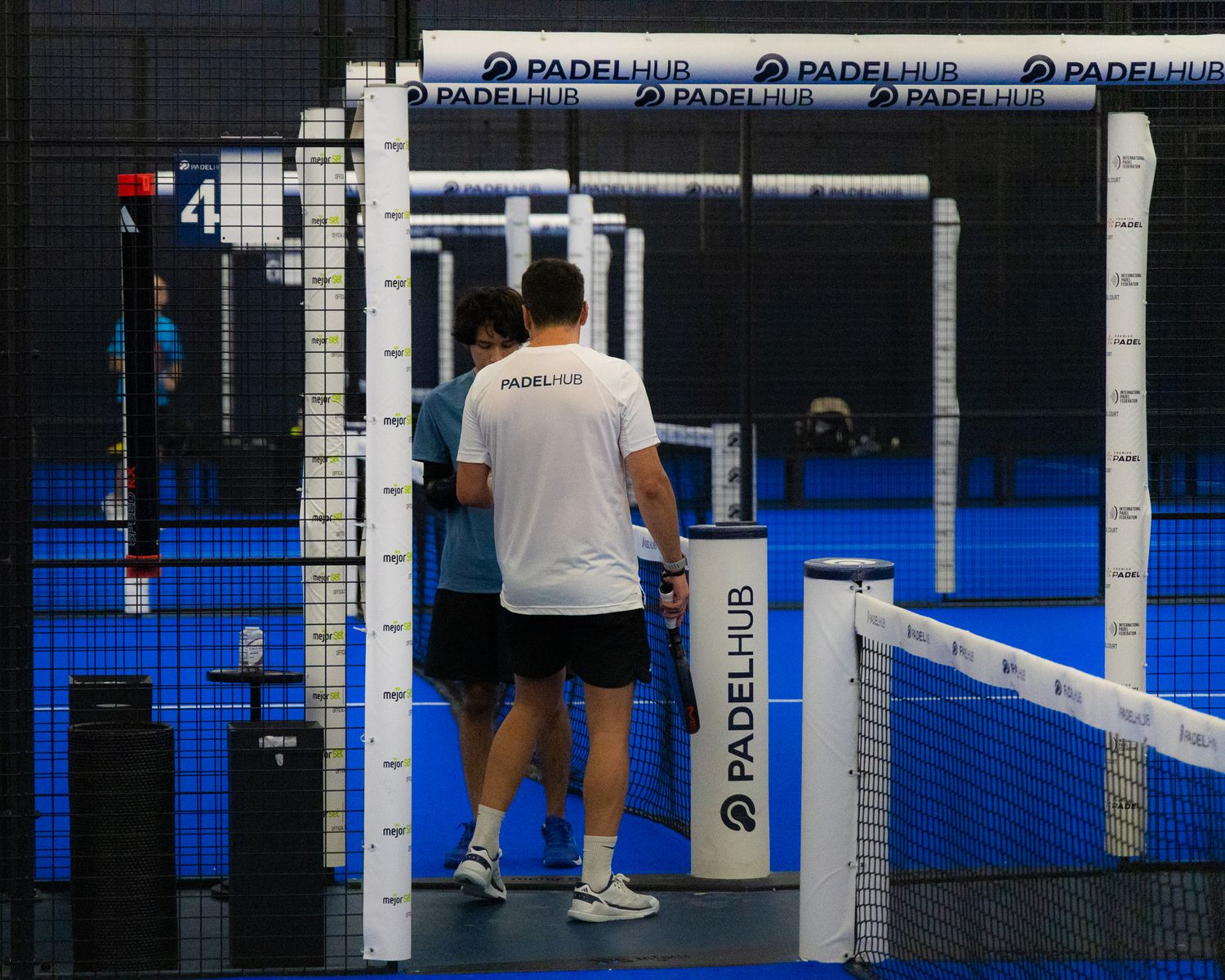 Two padel players standing at the net on a PadelHub branded indoor blue court.