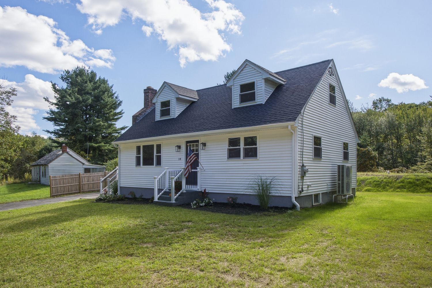 White Cape Cod home with dark roof, dormers, front porch, and lush green lawn surrounded by trees on a sunny day.