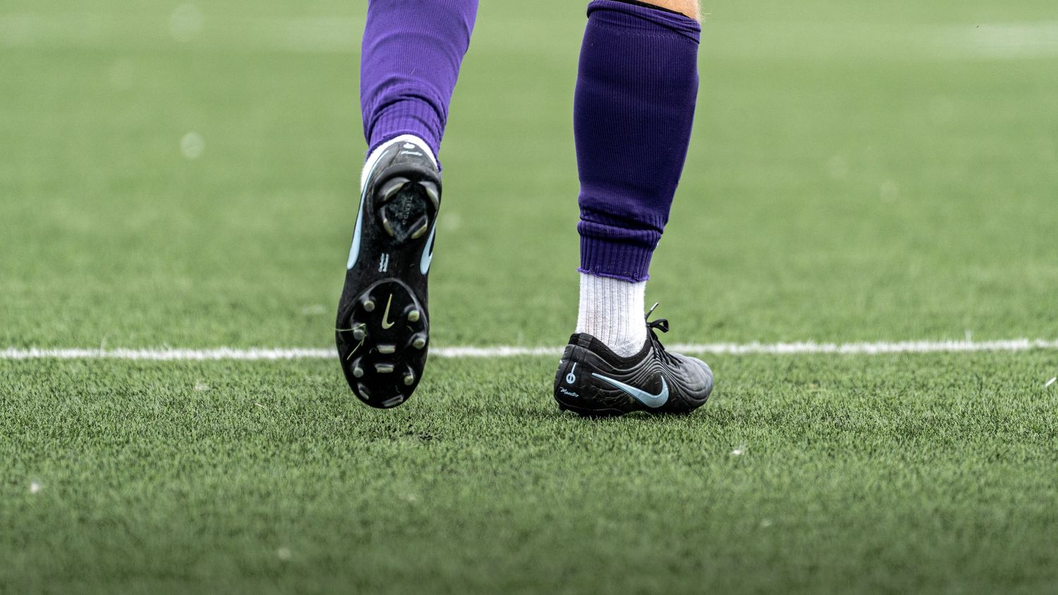 Close-up of soccer player wearing purple socks and black Nike cleats standing on green turf field.