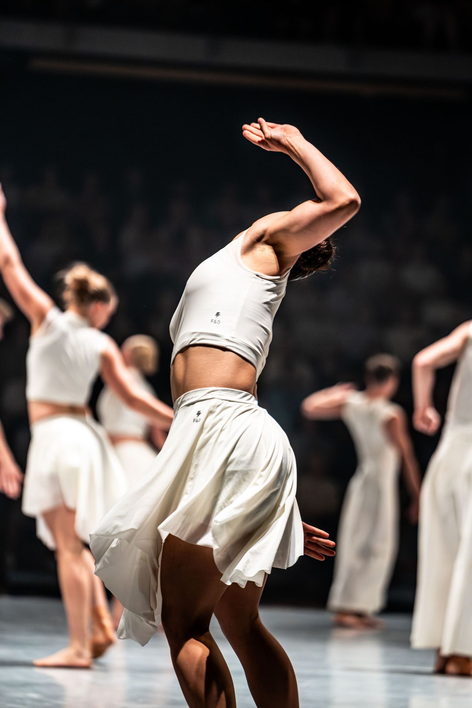 Female contemporary dancer in white crop top and skirt performs expressive back arch on stage.