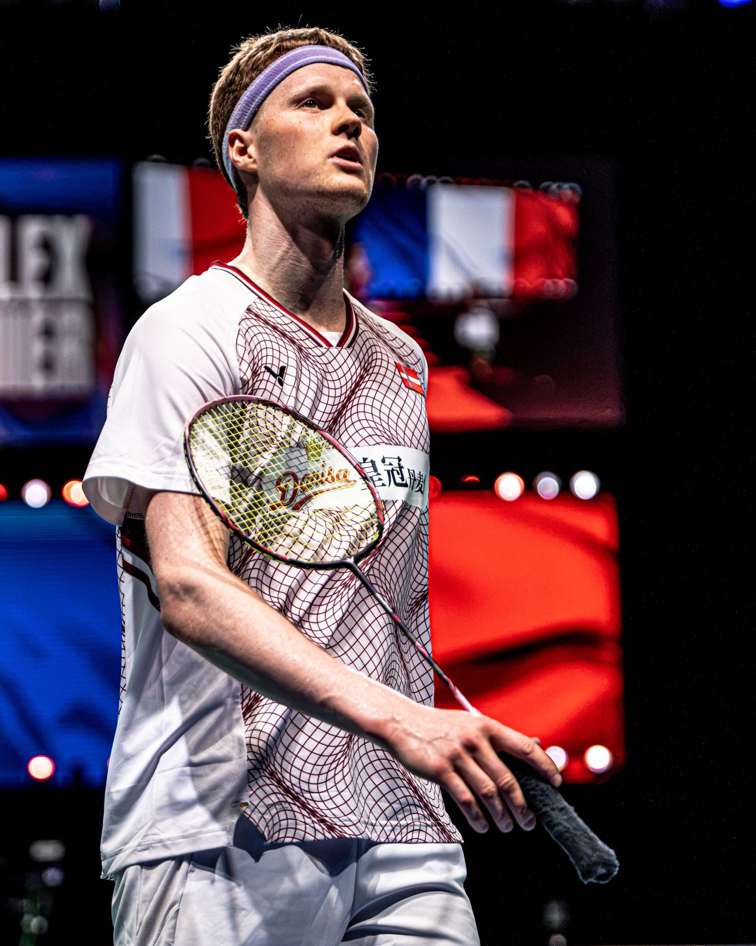 Anders Antonsen Danish badminton player holding racket with French flag visible on large screen in background.
