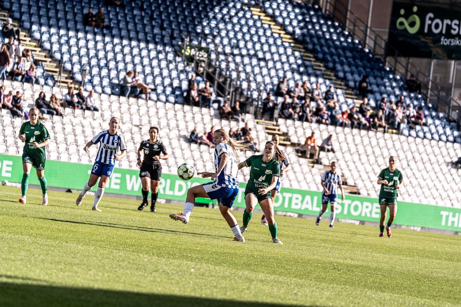 Female soccer player performs a bicycle kick during a match between blue-white and green-uniformed teams.