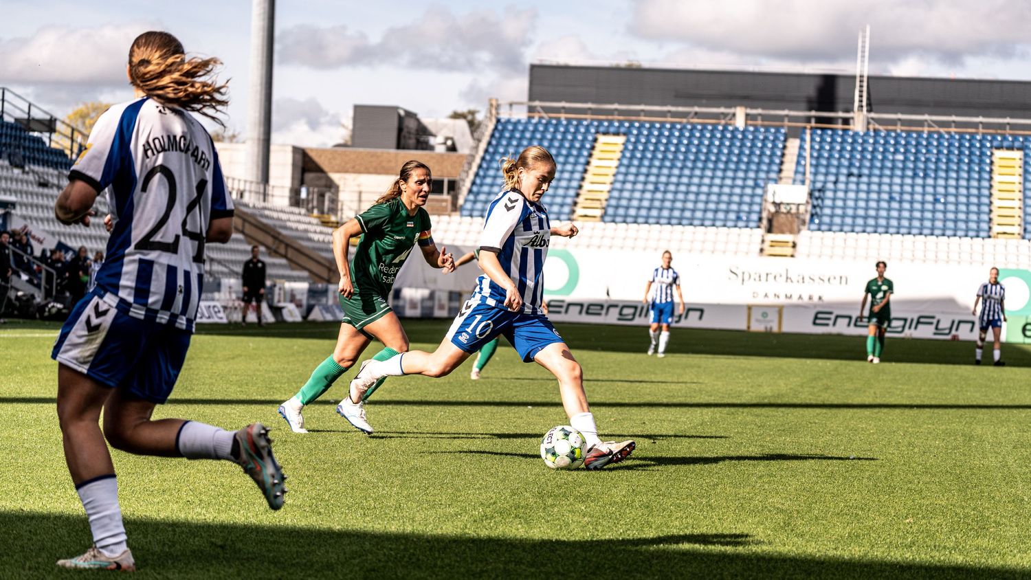 Female footballer in blue-white strip advances with ball during women's soccer match on grass pitch.