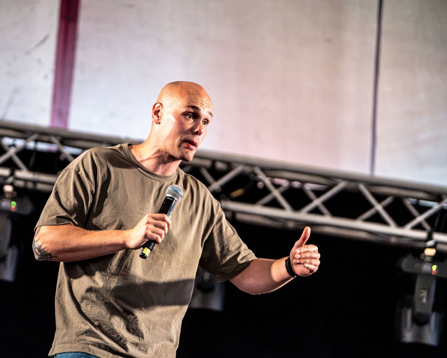 Bald man gesturing with a thumbs up while holding a microphone on stage.