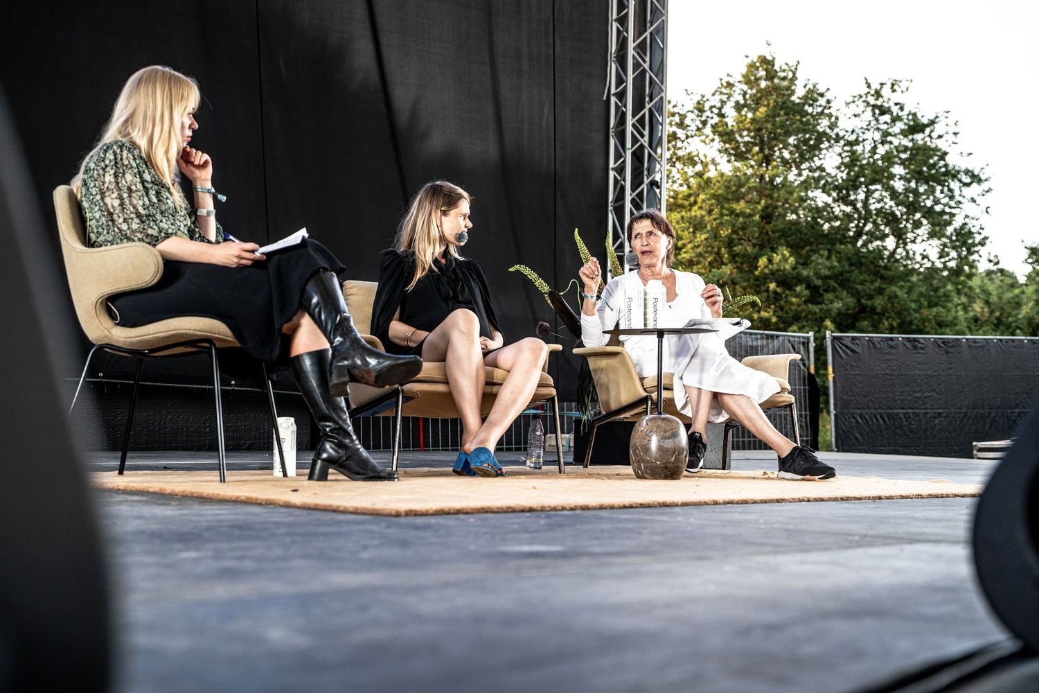 Three women participating in an outdoor panel discussion on a stage with lush greenery.
