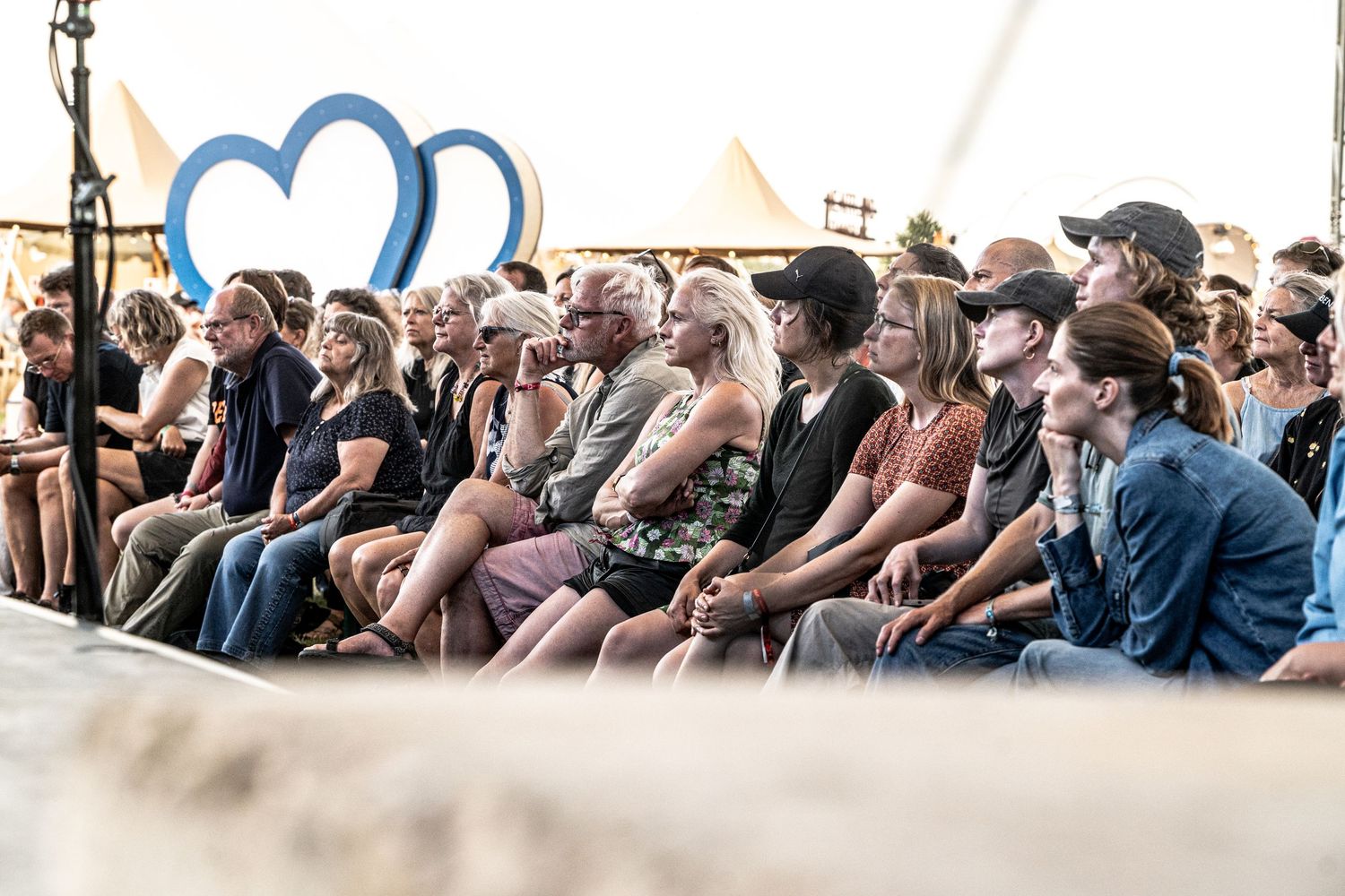 Large audience seated in front of a blue heart decoration at an outdoor festival stage.