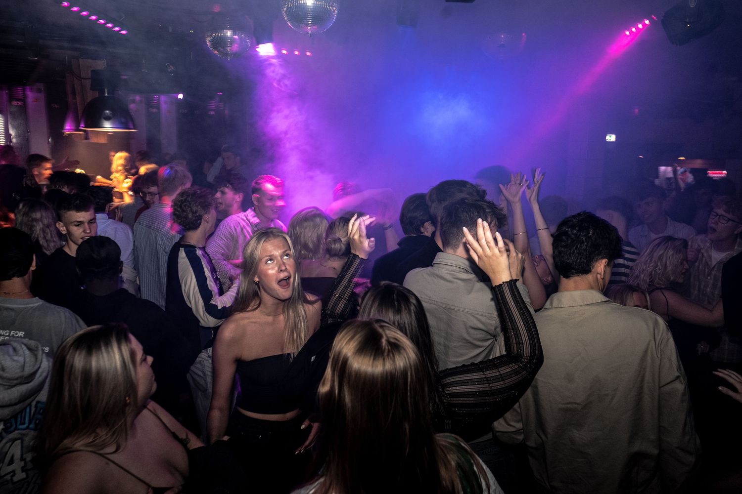 Packed nightclub dance floor with partygoers dancing and cheering under purple and pink stage lights and haze.
