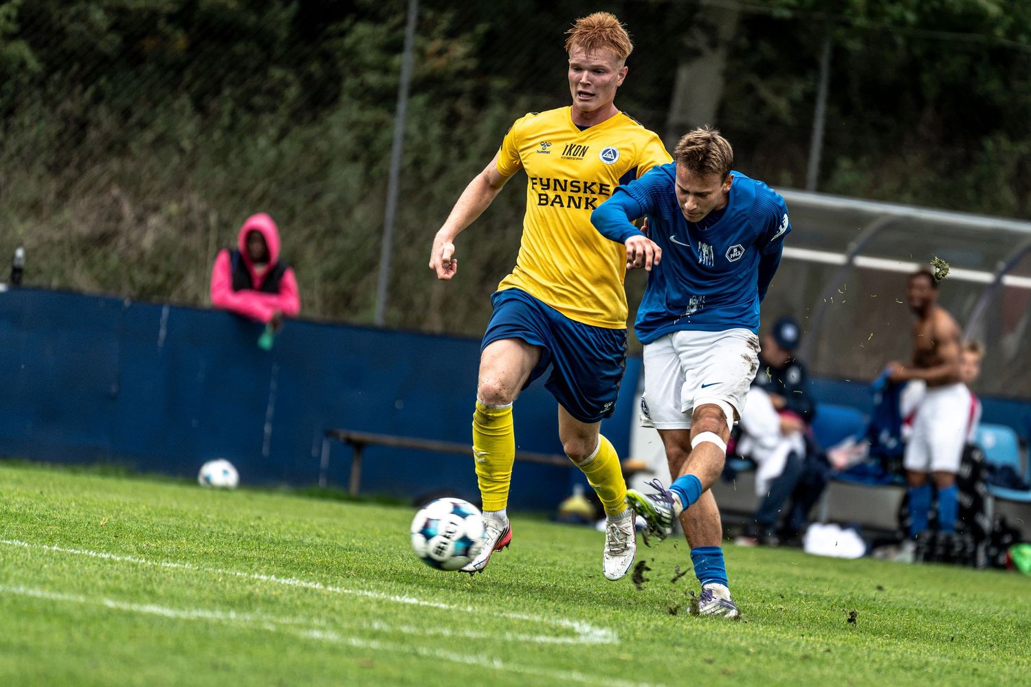 Two soccer players compete for the ball during a match, one in yellow and one in blue uniforms, on a green field.