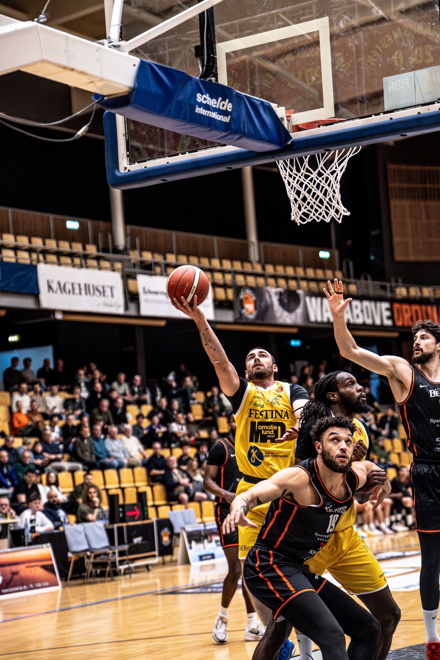 Yellow team player drives to the basket past defenders in an indoor basketball game.