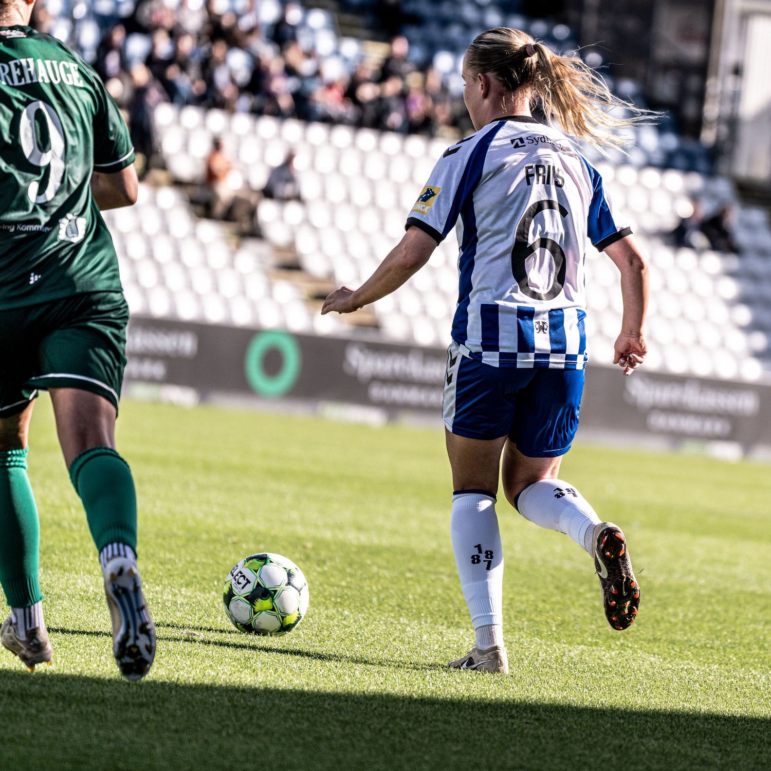 Female soccer player in blue and white dribbles past a green-uniformed defender during a competitive match.