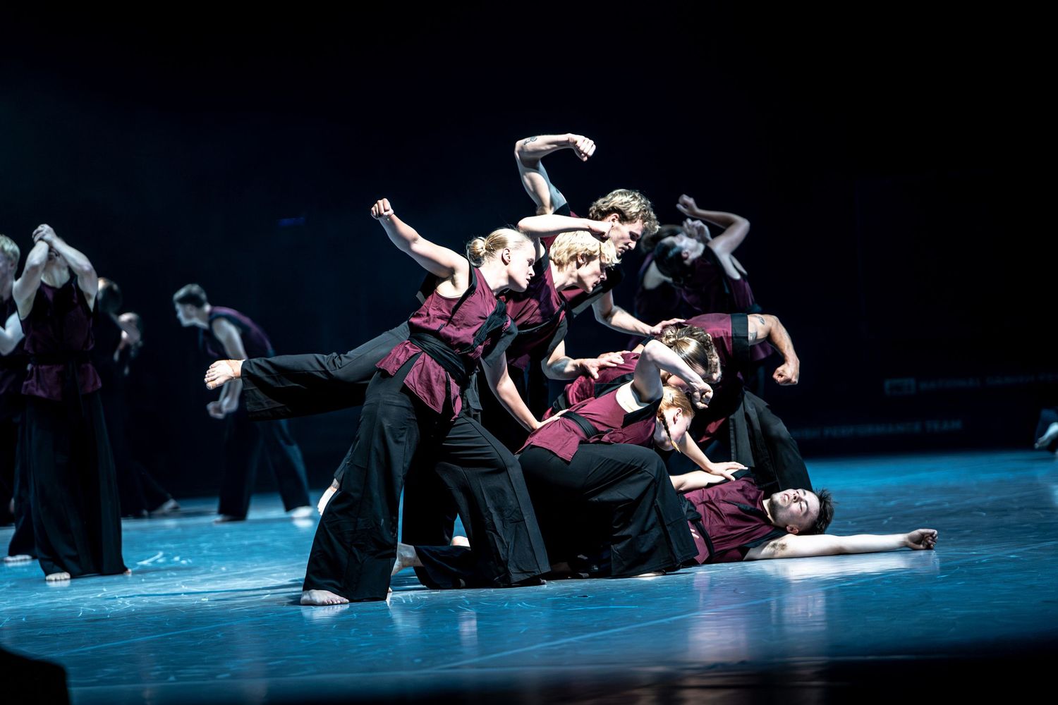 Large ensemble of dancers in black and burgundy costumes performing a dynamic group formation.