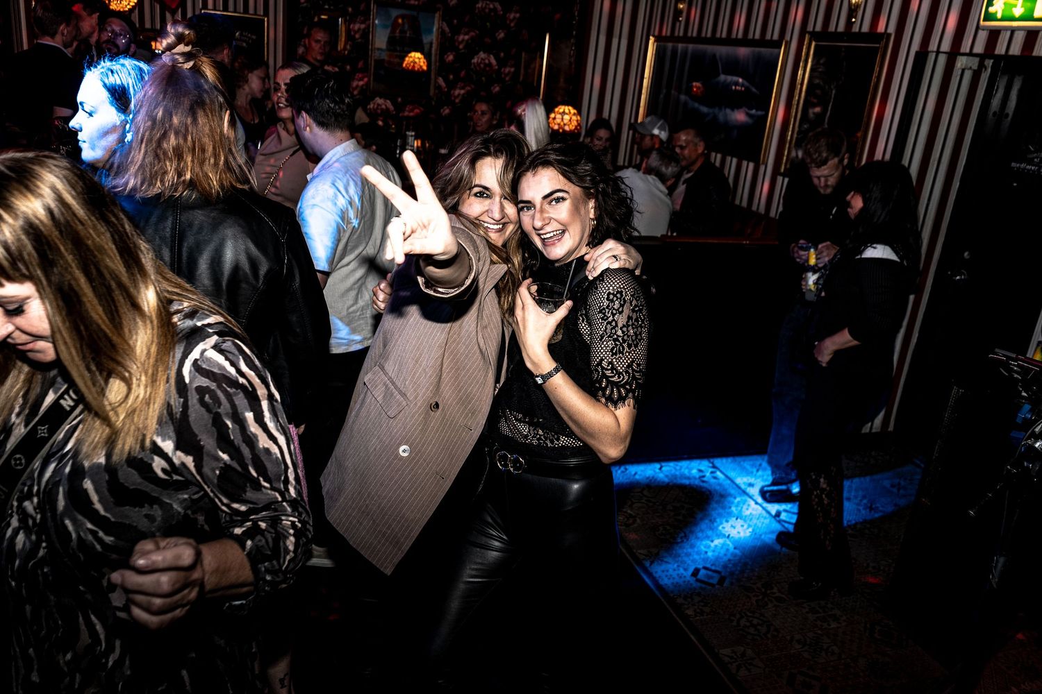 Two smiling women pose together and flash peace signs on a crowded nightclub dance floor lit by blue lights.