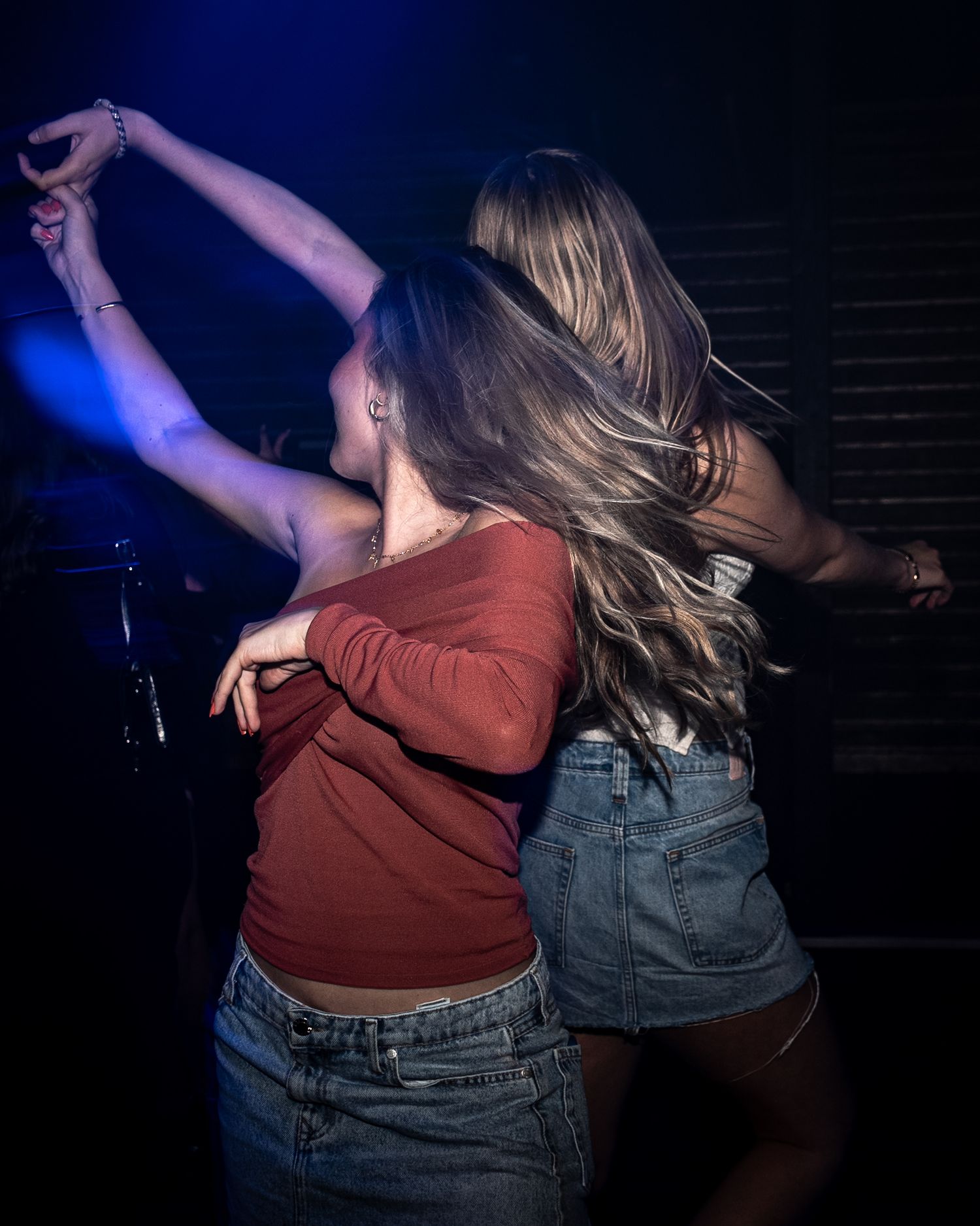 Two women with long hair dance together in a dimly lit club, one in a red top and denim skirt, under blue stage lighting.
