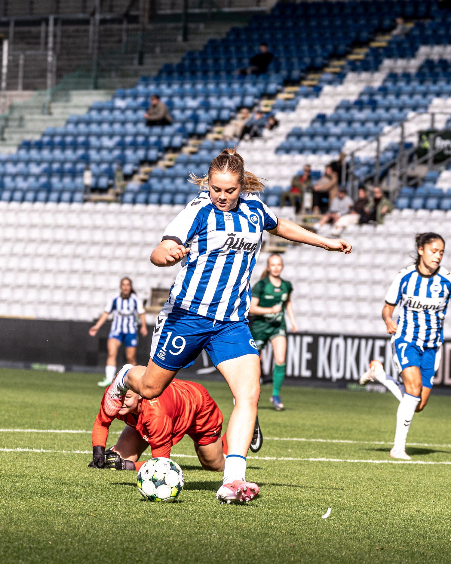 Female soccer player number 19 in blue-white kit dribbles past fallen goalkeeper during match.