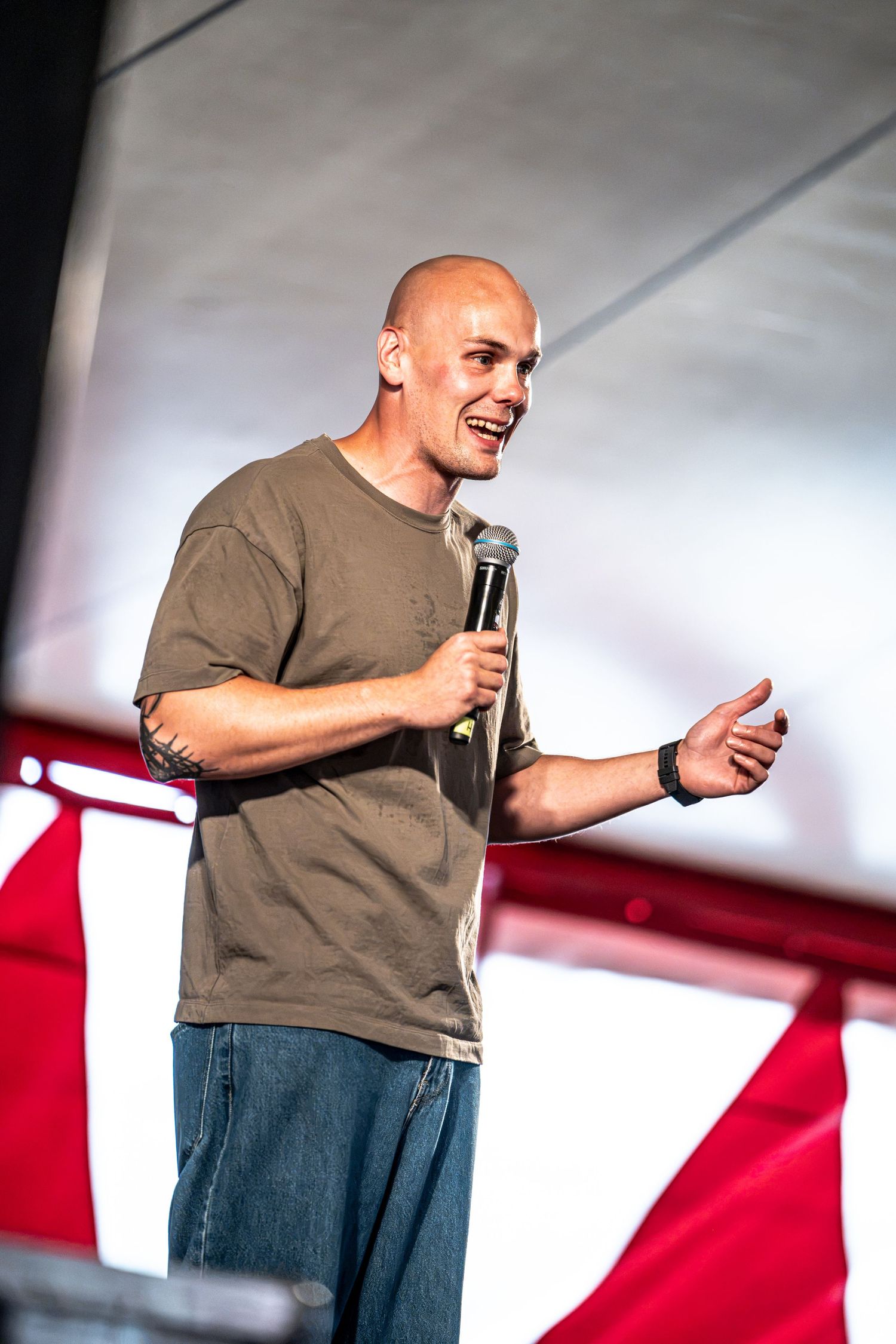 Bald man in olive shirt performing on stage with a microphone and thumbs up.