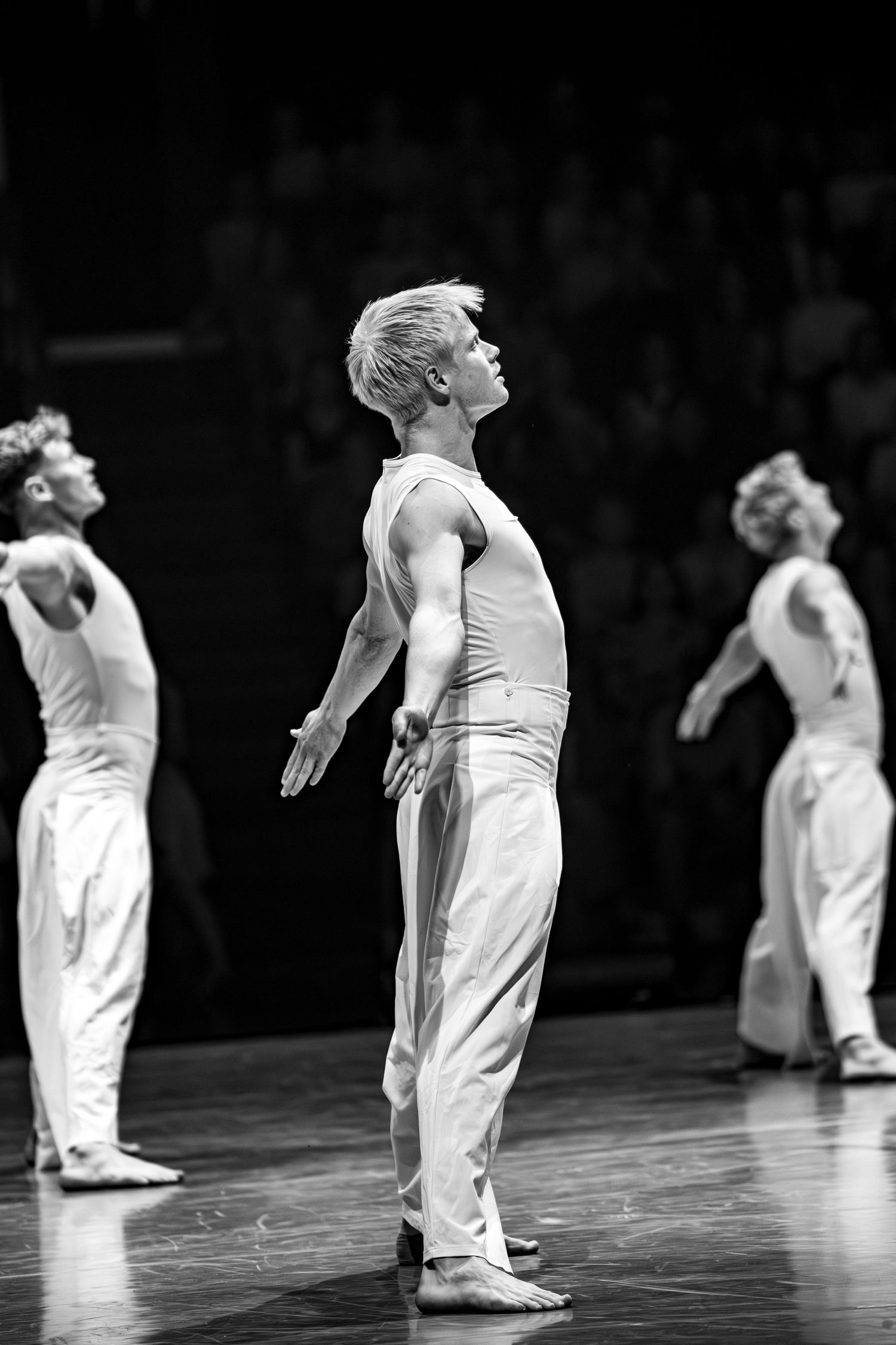 Male dancer in white costume gazes upward gracefully on stage in black and white photo.