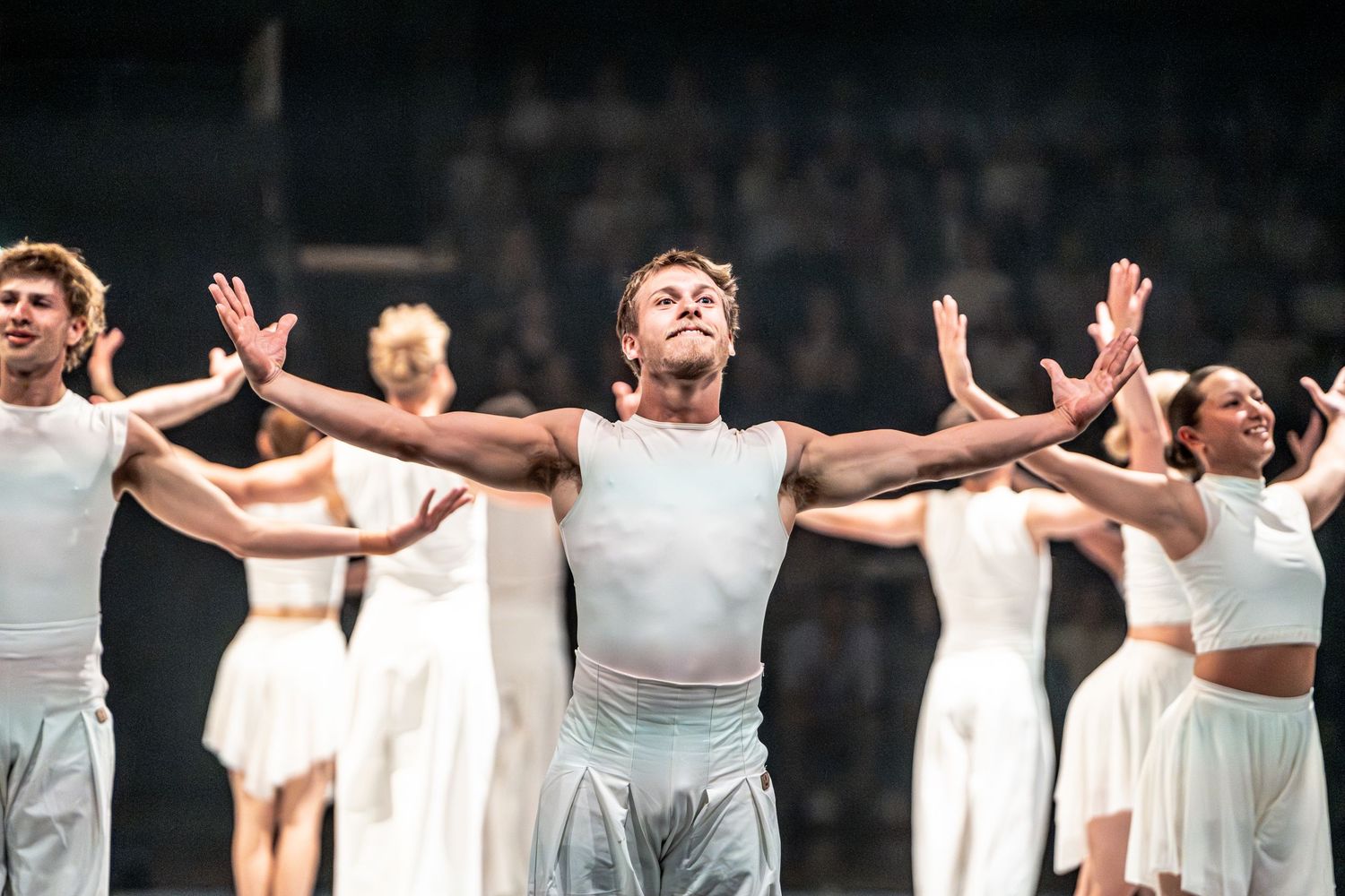 Male dancer in white costume stands center stage arms outstretched surrounded by ensemble performers.