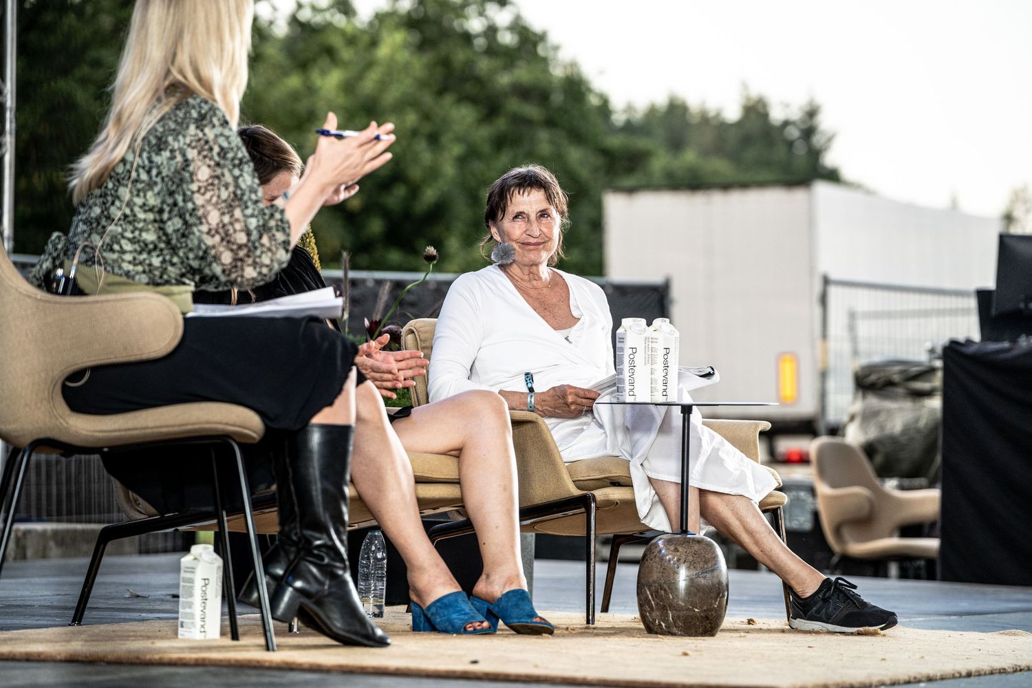 Two women in relaxed conversation on an outdoor stage, one in white dress smiling warmly.