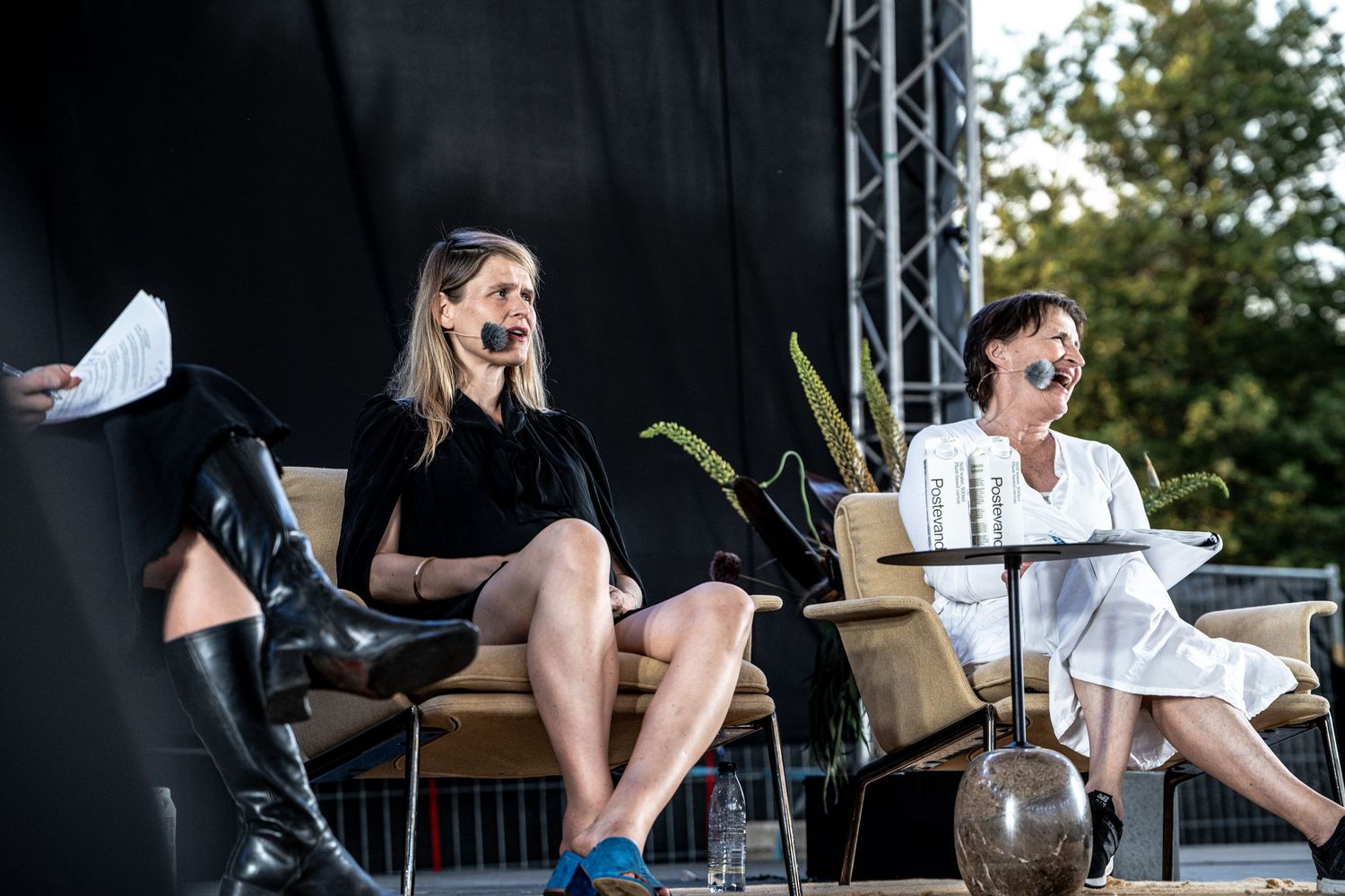 Two women seated on stage outdoors during a panel discussion, one in black dress and one in white outfit.