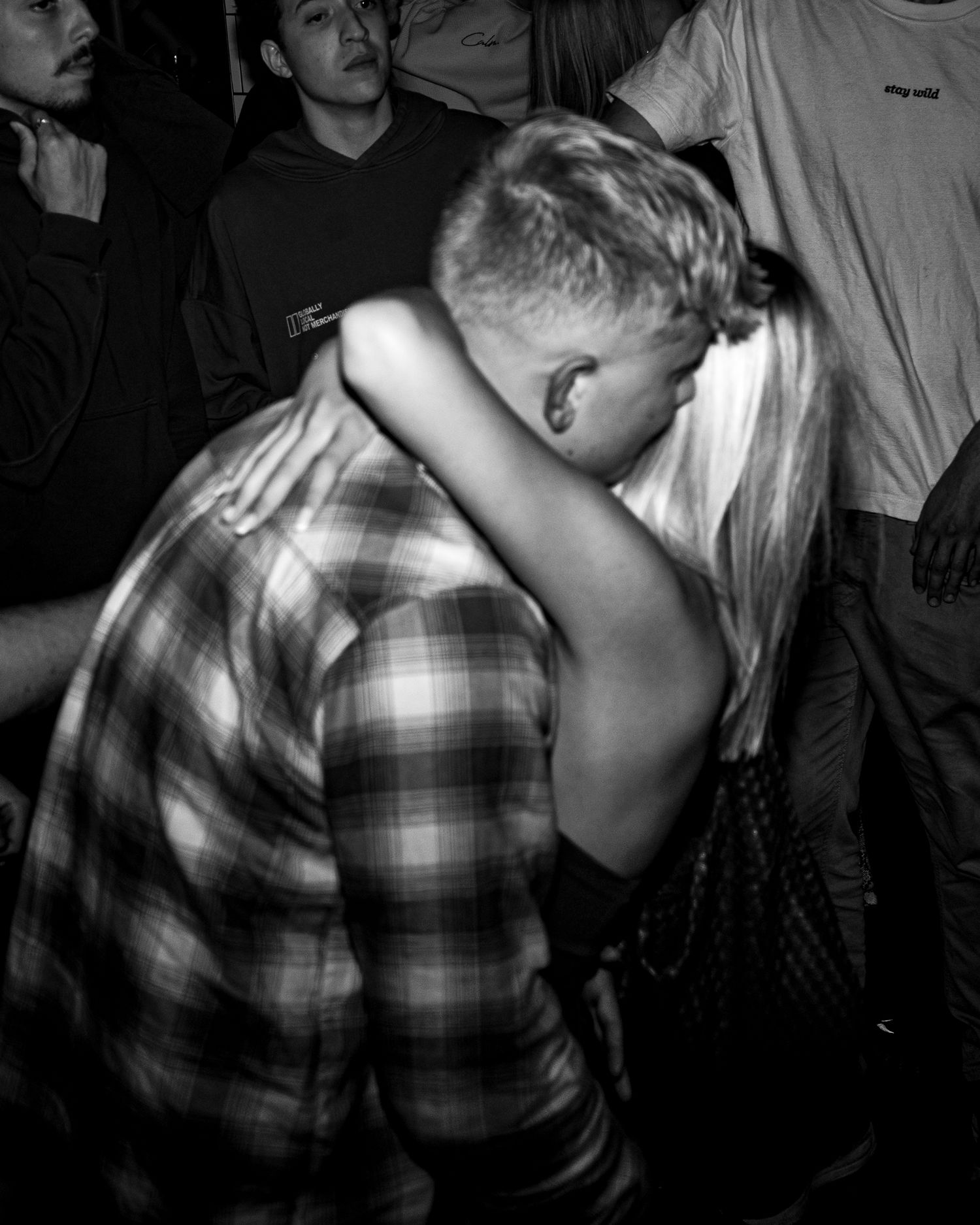 Black and white photo of a couple embracing in a crowded nightclub, with onlookers in hoodies and t-shirts behind them.