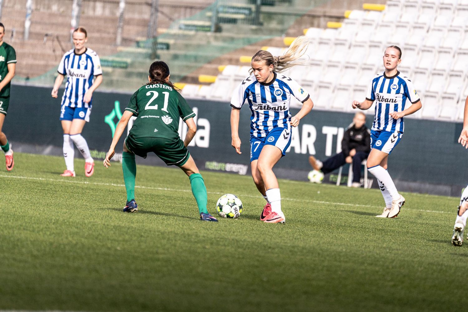 Female soccer player in blue and white kit dribbling past opponent number 21 in green during league match.