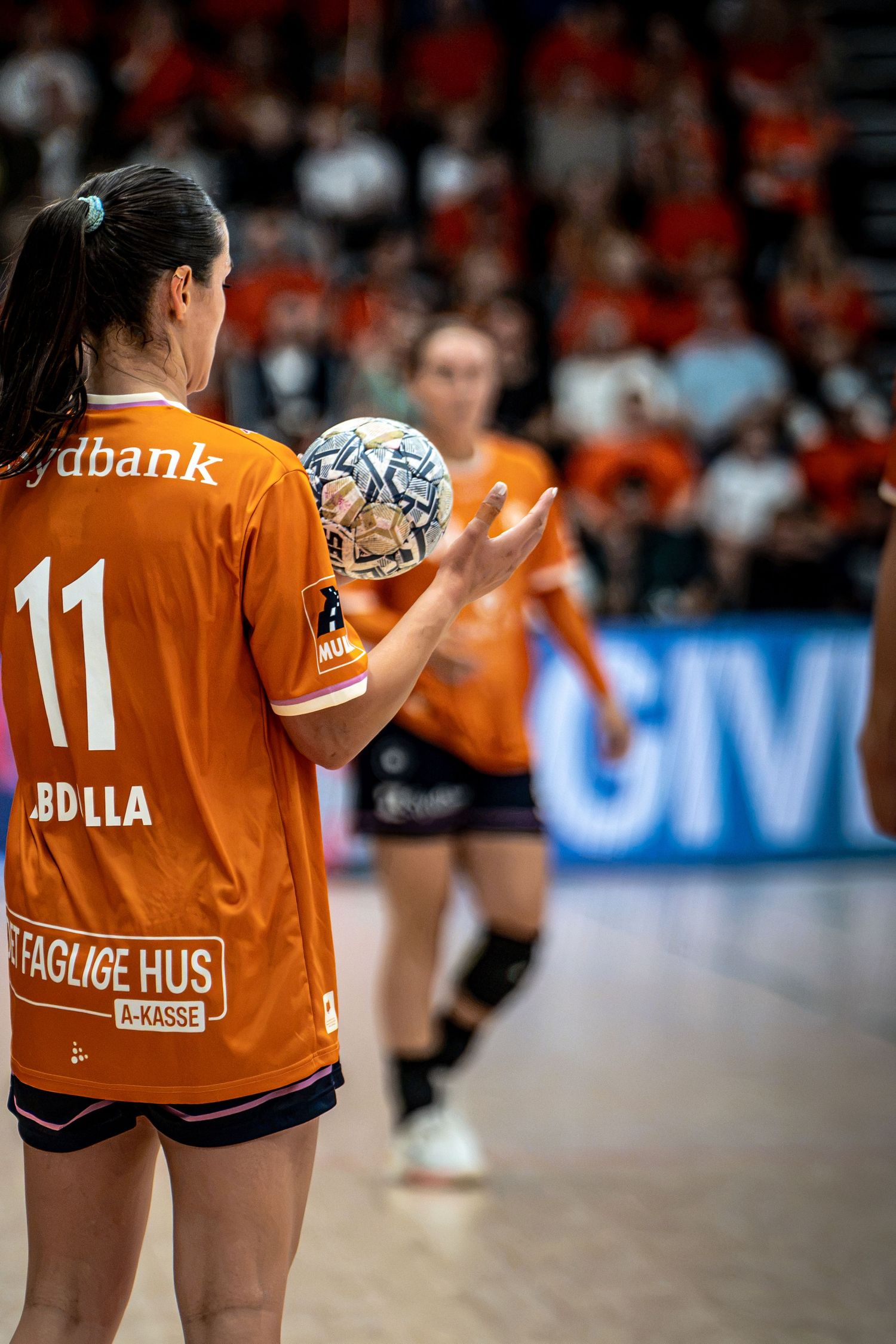 Female handball player number 11 holds ball during women's indoor handball match before orange crowd.