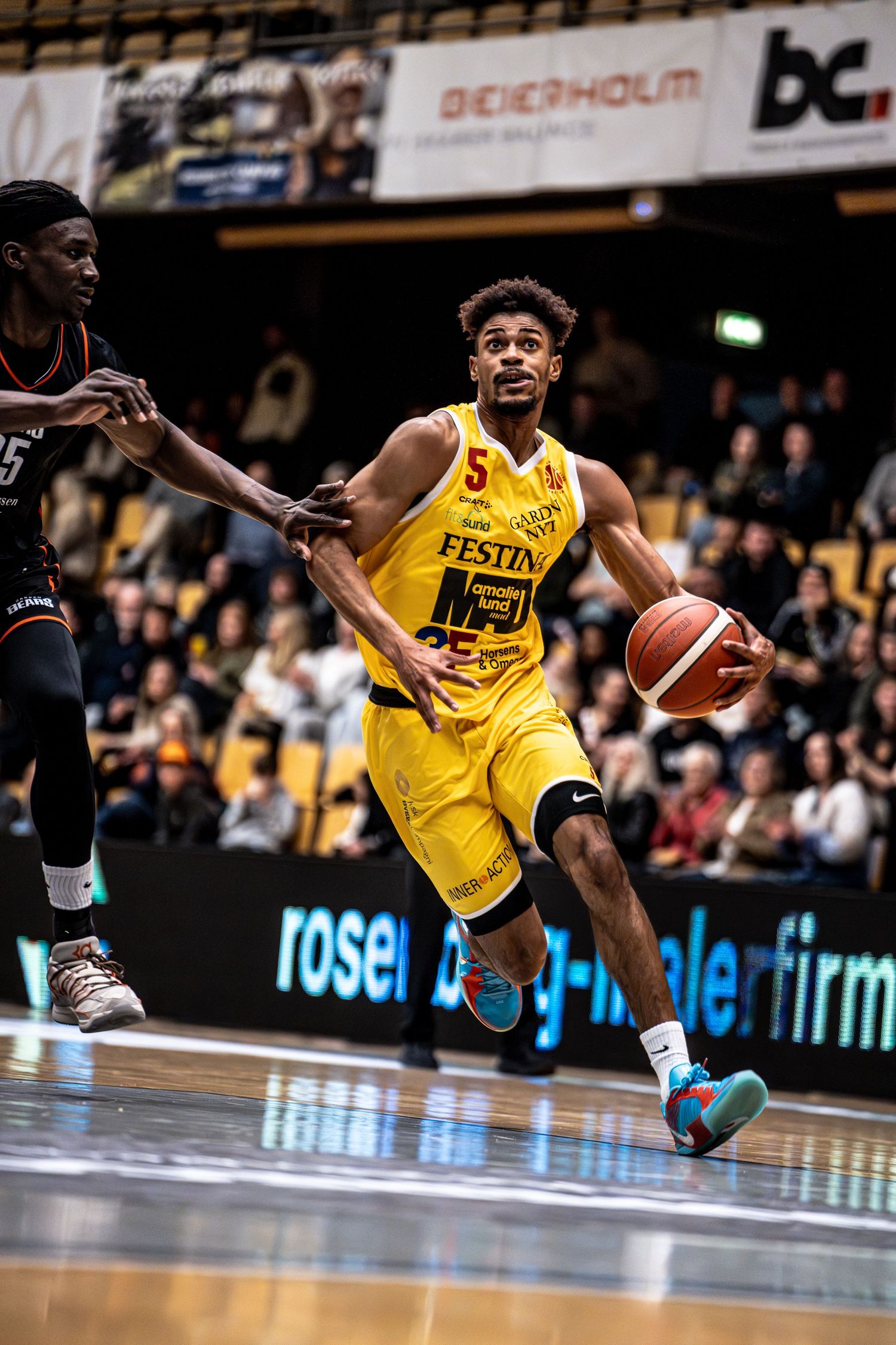 Yellow-uniformed player number 5 drives baseline past a black-uniformed defender indoors.