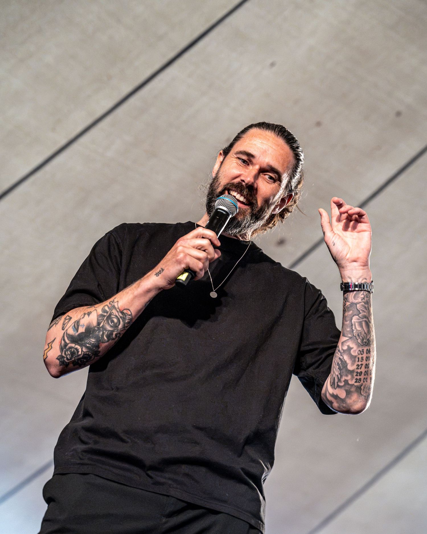 Bearded long-haired tattooed male vocalist performing on stage in a black t-shirt with headphones around neck.