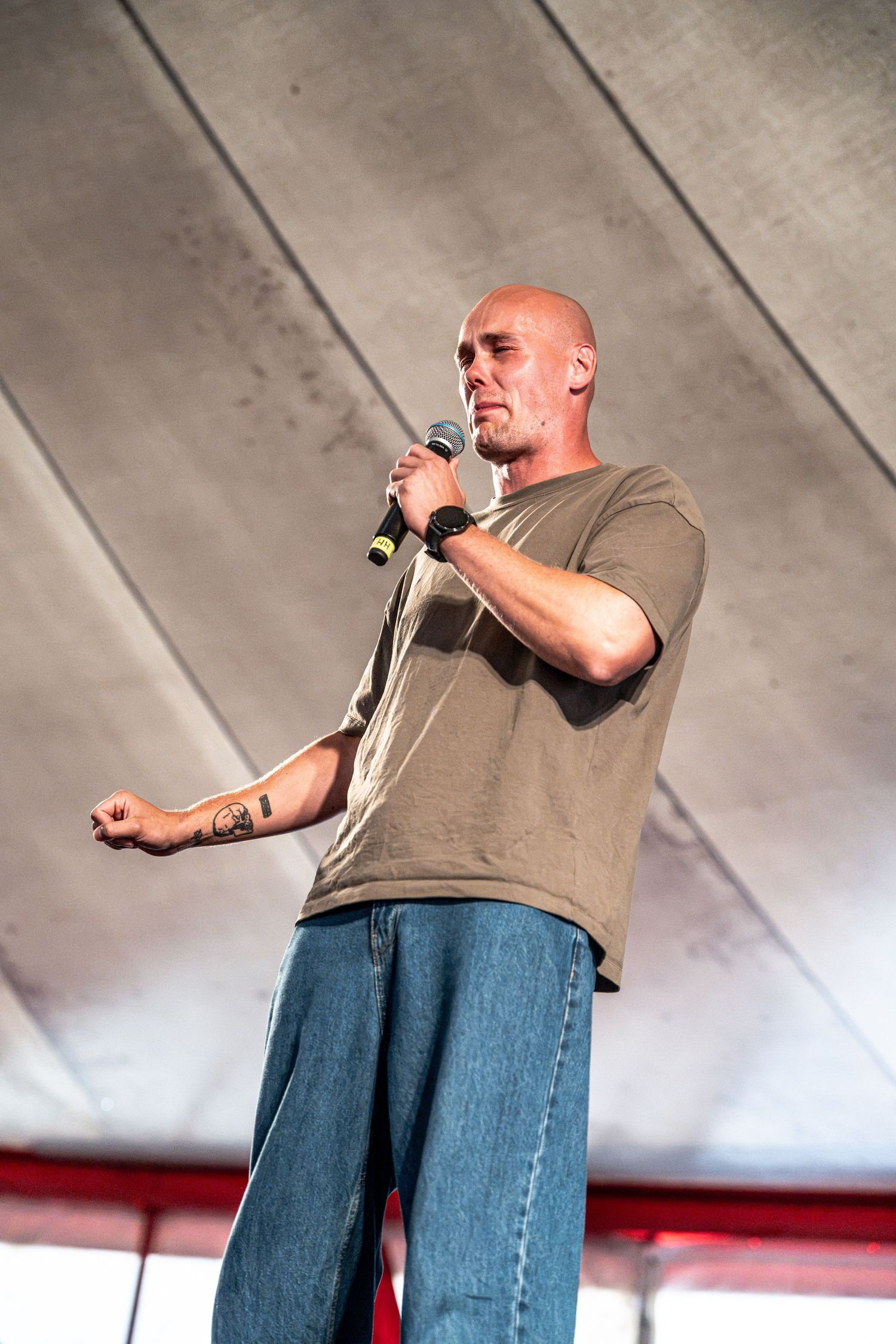 Bald male performer in olive t-shirt and jeans sings into microphone on an outdoor festival stage.