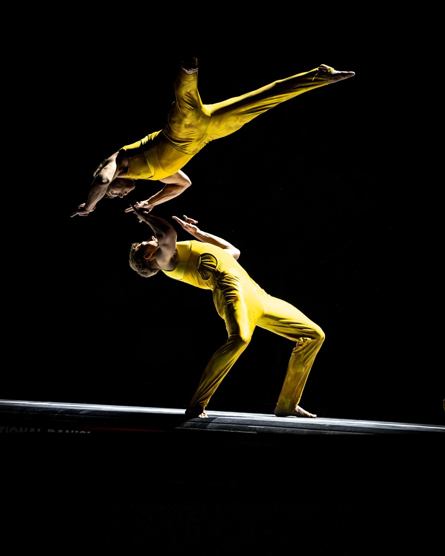 Two acrobats in yellow suits perform stunning balance act on dark dramatic stage.