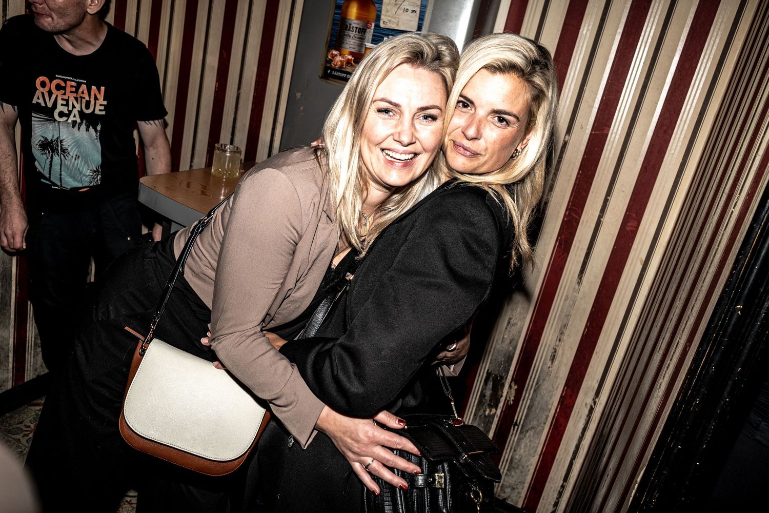 Two smiling blonde women in dark jackets embrace and pose together at a lively bar while a man in a graphic t-shirt stands behind them.