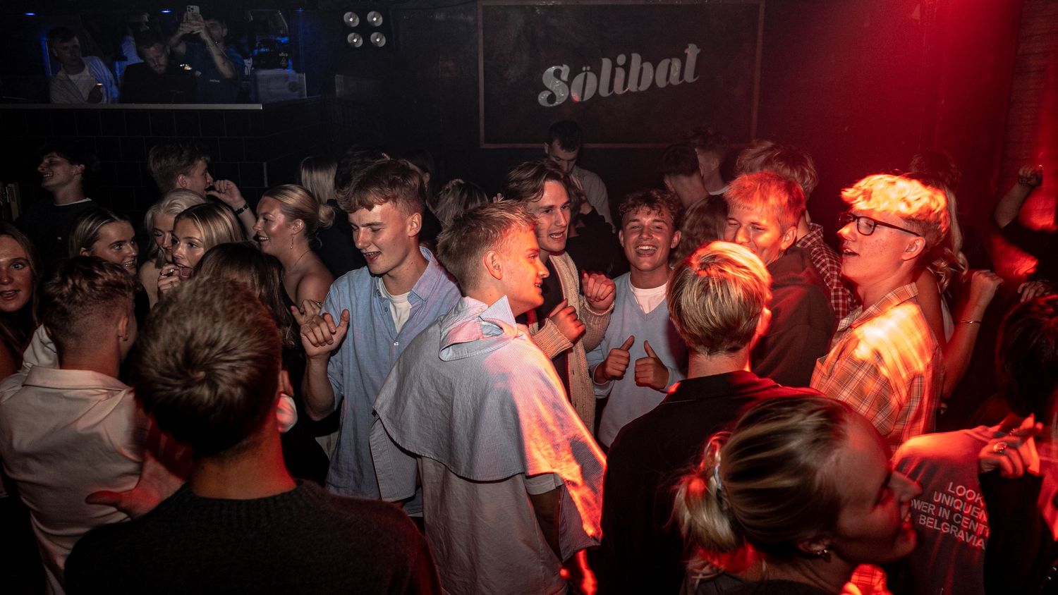 Crowd of cheering young partygoers dancing and raising their hands in a red-lit club beneath a Solibal sign.