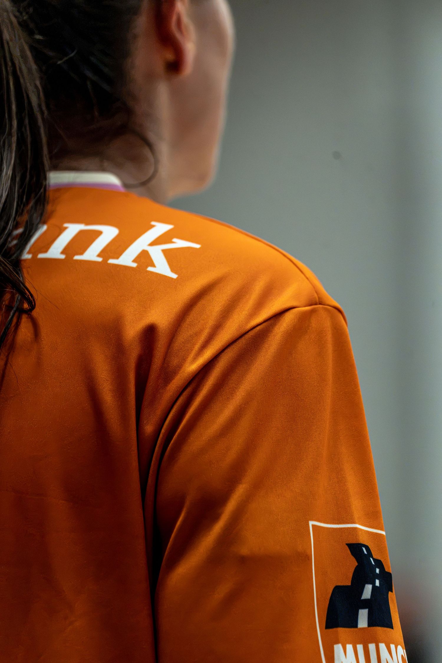 Close-up rear view of female athlete wearing orange handball jersey with sponsor logo visible.