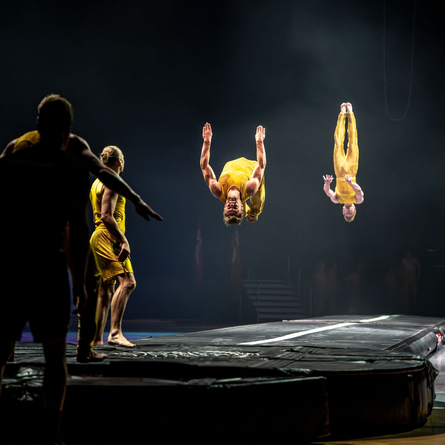 Acrobats in yellow suits performing backflips on a trampoline during a dramatic circus performance under stage lighting.