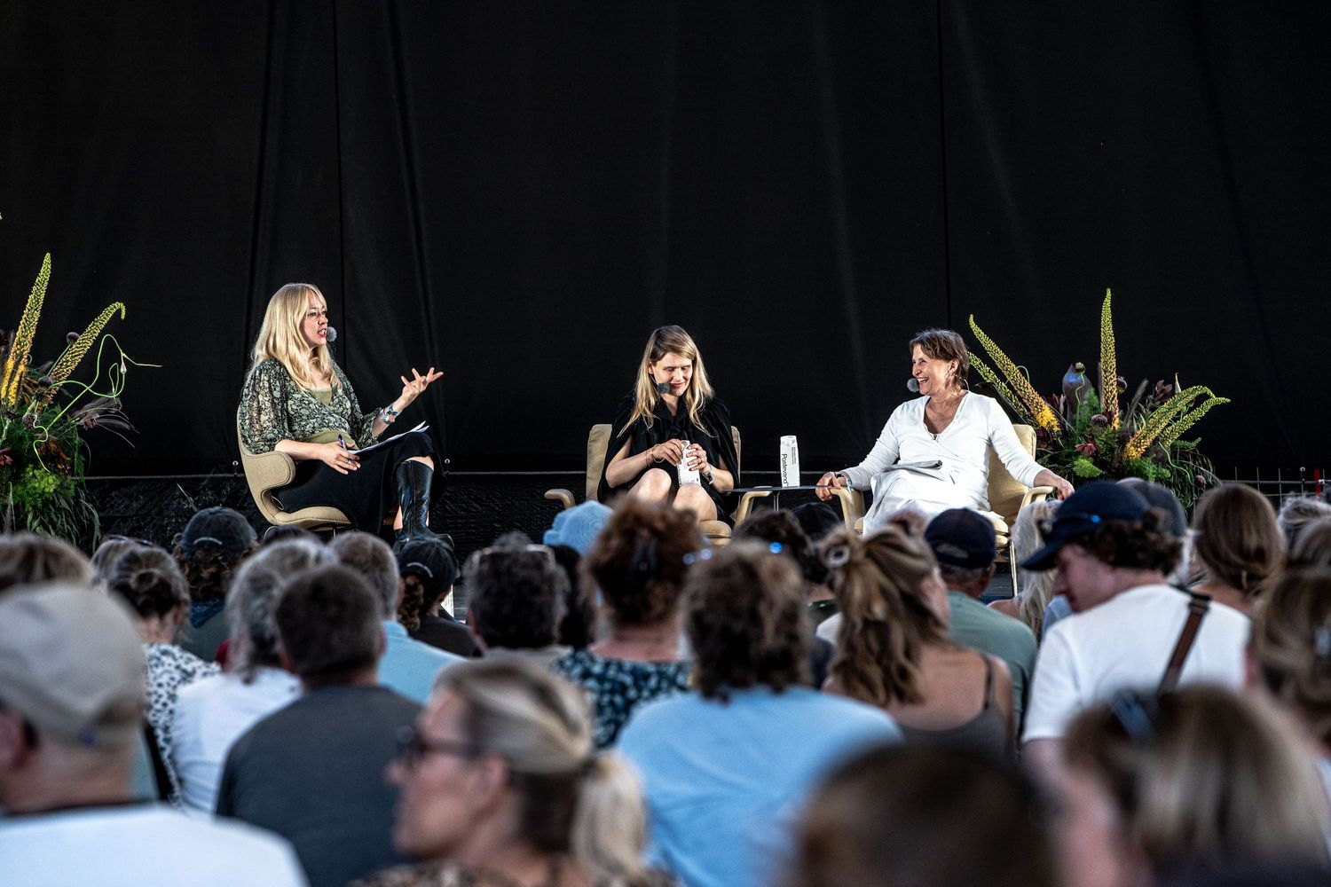 Three women in animated discussion on stage at outdoor festival.