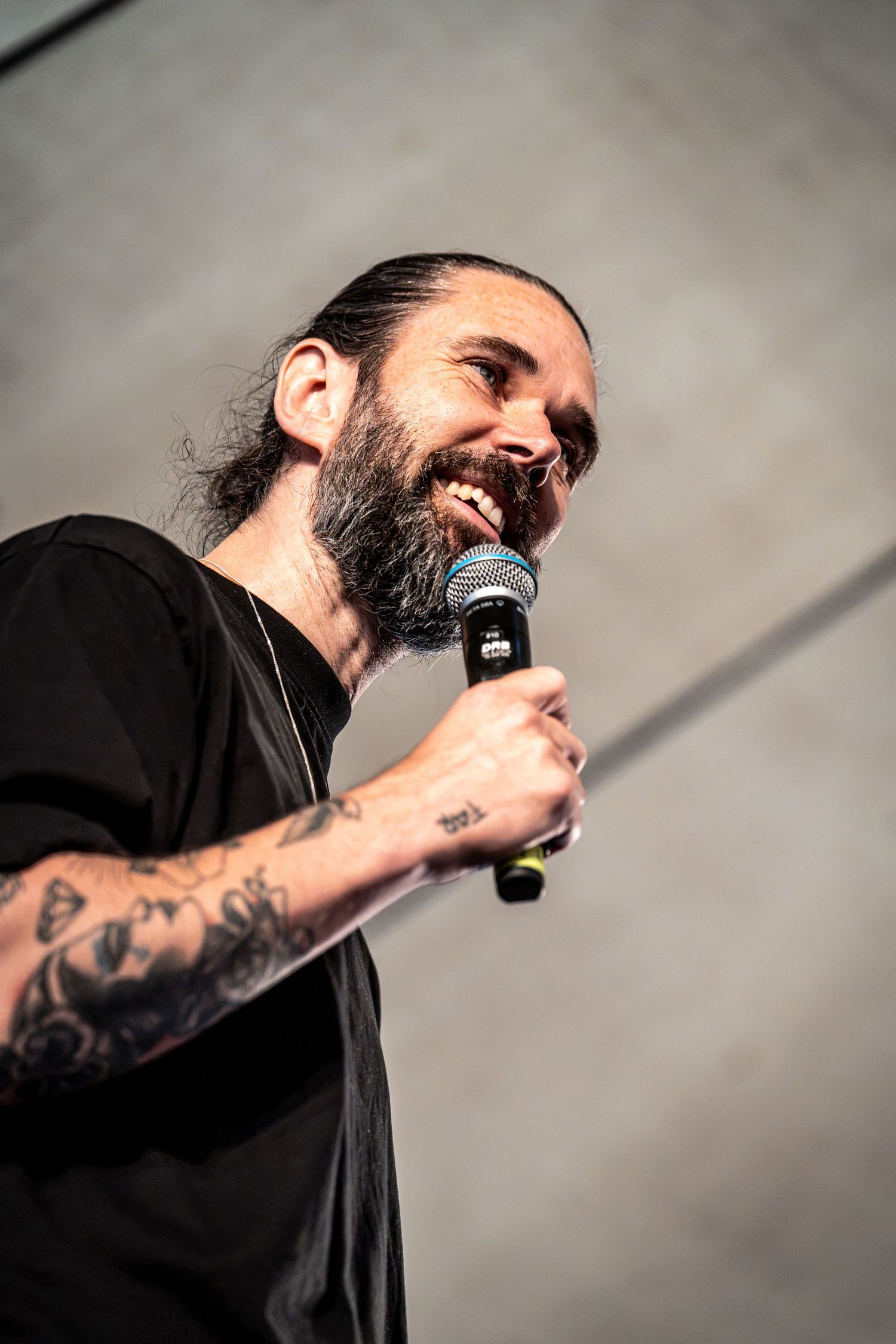 Smiling bearded performer with tattoos sings into microphone wearing black t-shirt on stage.