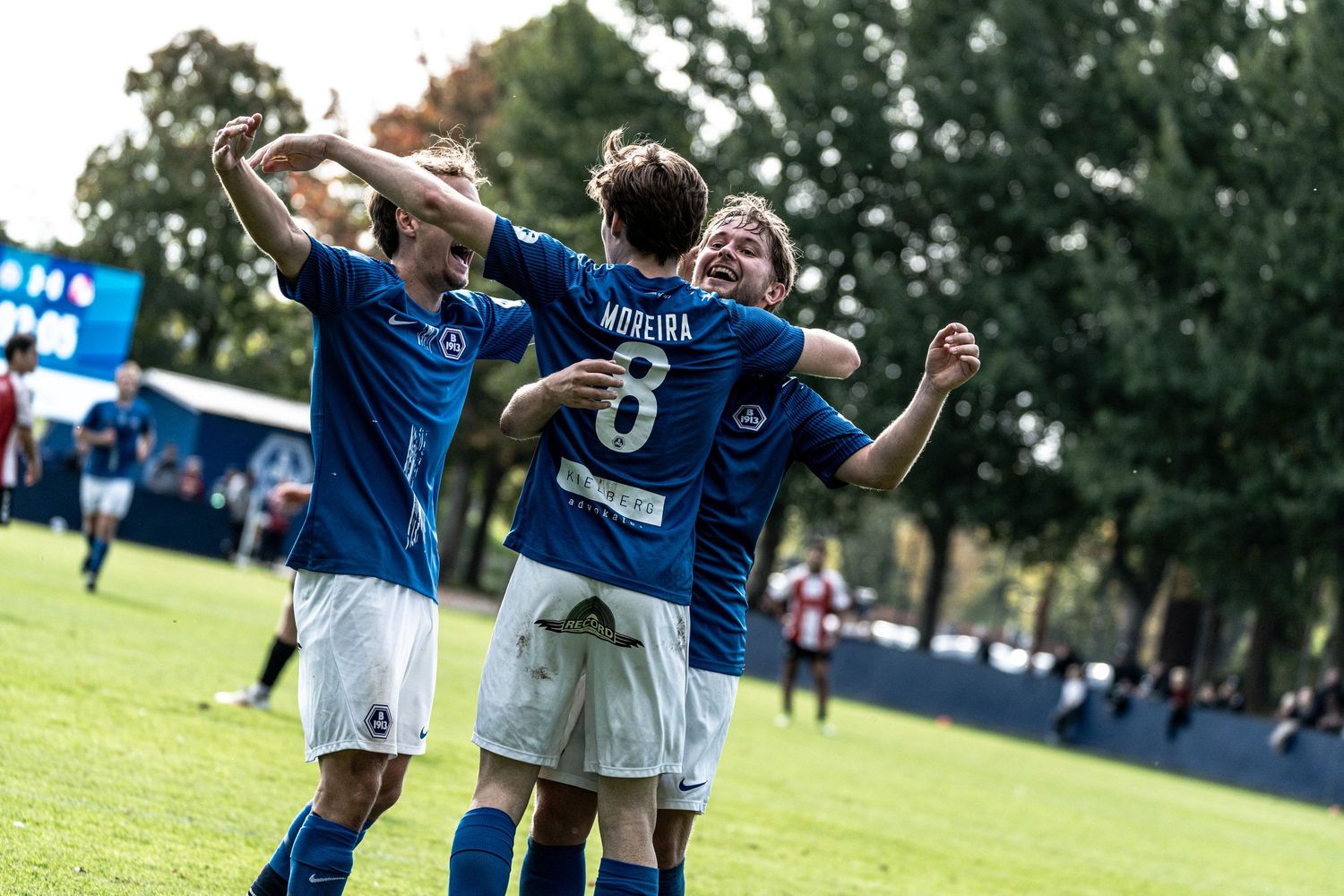 Soccer players in blue uniforms celebrate together on the field during a sunny outdoor match.