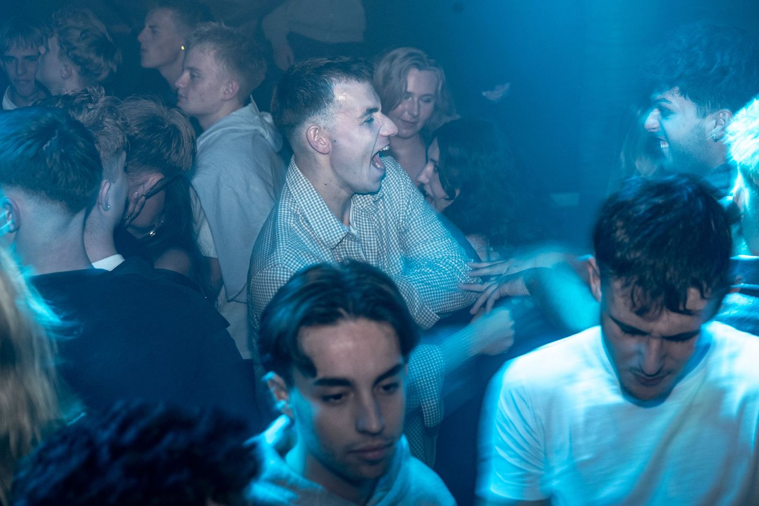 Crowd of young men packed together on a crowded nightclub dance floor, laughing and singing under blue club lighting.