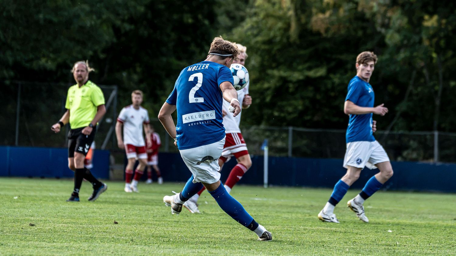 Soccer player number 2 in blue kit kicks ball during outdoor match with referee and teammates visible.