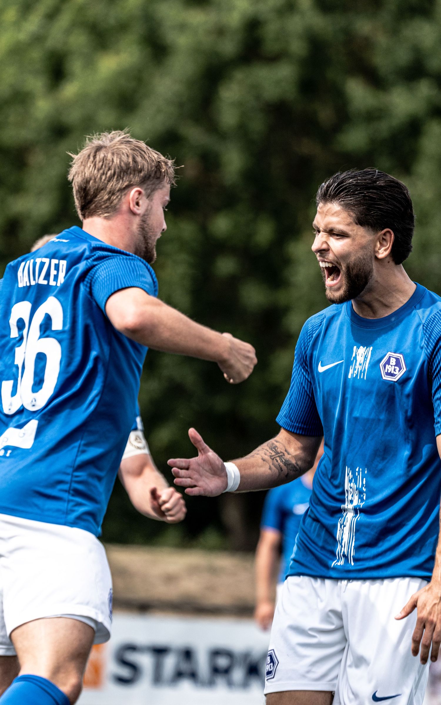 Two soccer players in blue Nike uniforms celebrate a goal, with player number 36 named Kruzer shaking hands.